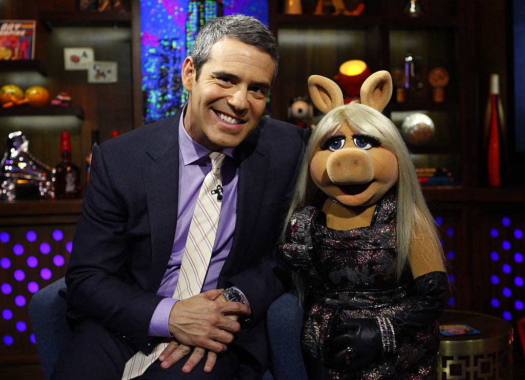 Andy Cohen, in a suit and tie, sitting next to Miss Piggy, wearing an elegant dress, on a TV show set