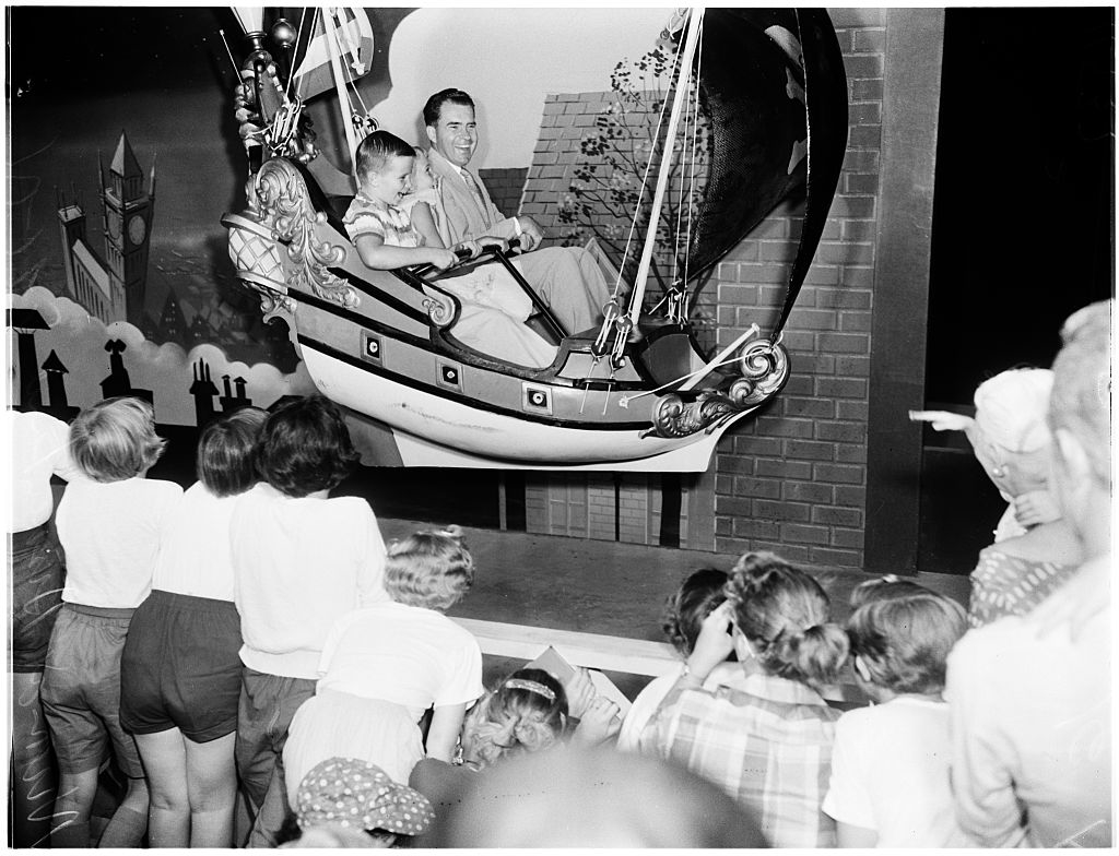 Walt Disney and a young girl riding a Peter Pan&#x27;s Flight attraction as a crowd of children watches in awe
