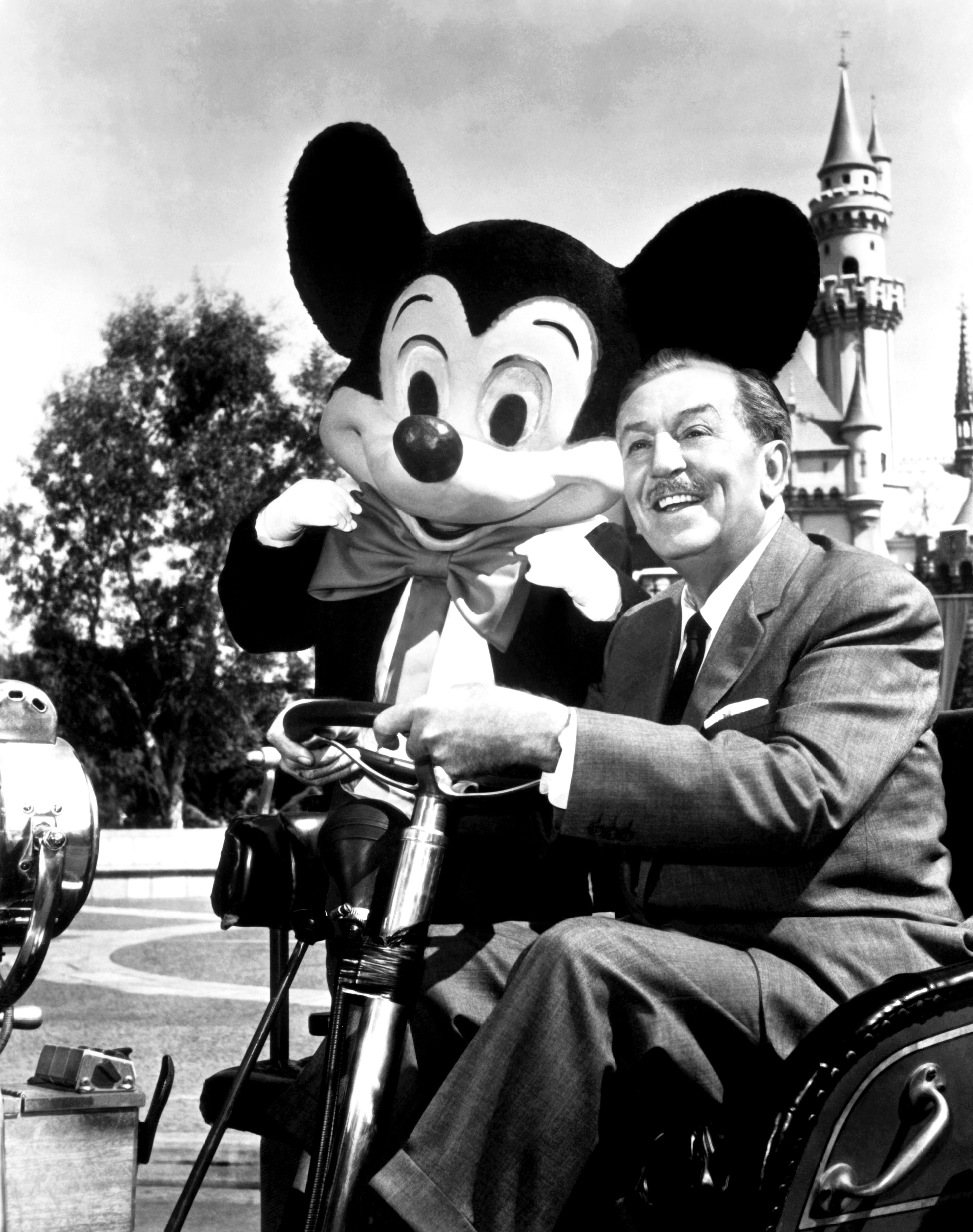 Walt Disney and a person in a Mickey Mouse costume at Disneyland, with Sleeping Beauty Castle in the background. Disney is seated and smiling