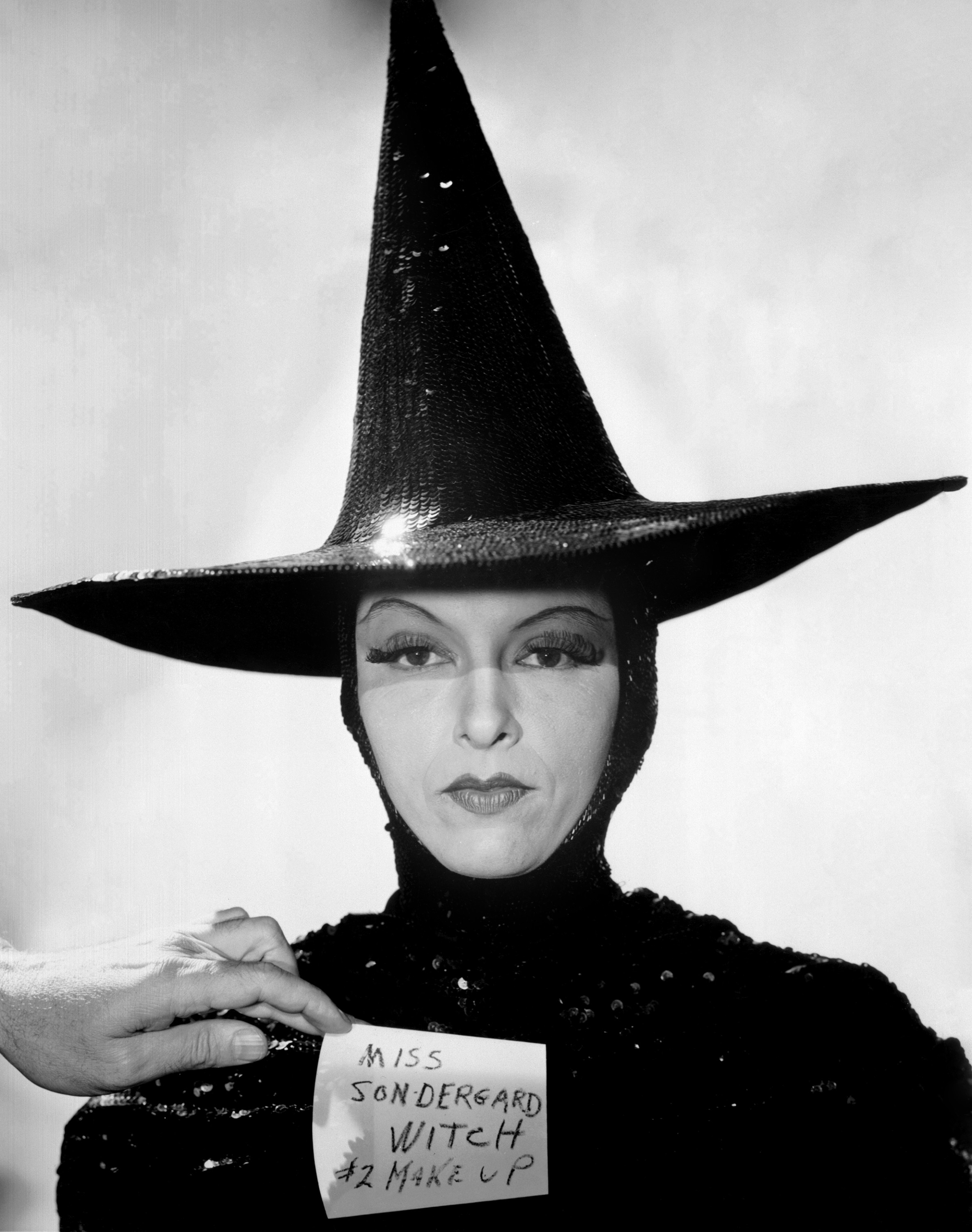 Gale Sondergaard dressed as a witch, wearing a high pointed hat and black glittering attire, holding a card that says &quot;Miss Sondergard Witch #2 Make Up.&quot;