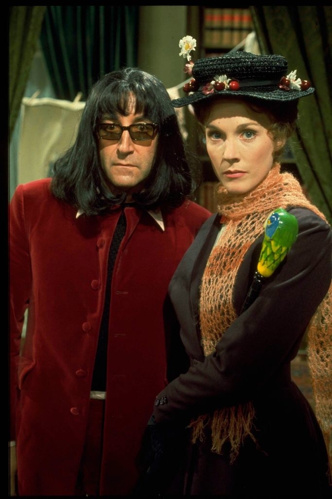 Two people in costumes. One in a red velvet suit and dark sunglasses, the other dressed as Mary Poppins with a parrot umbrella