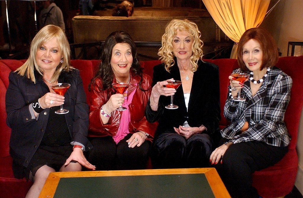 Four women are seated on a couch, smiling and holding cocktail glasses. They are dressed in stylish clothing, including jackets and blouses