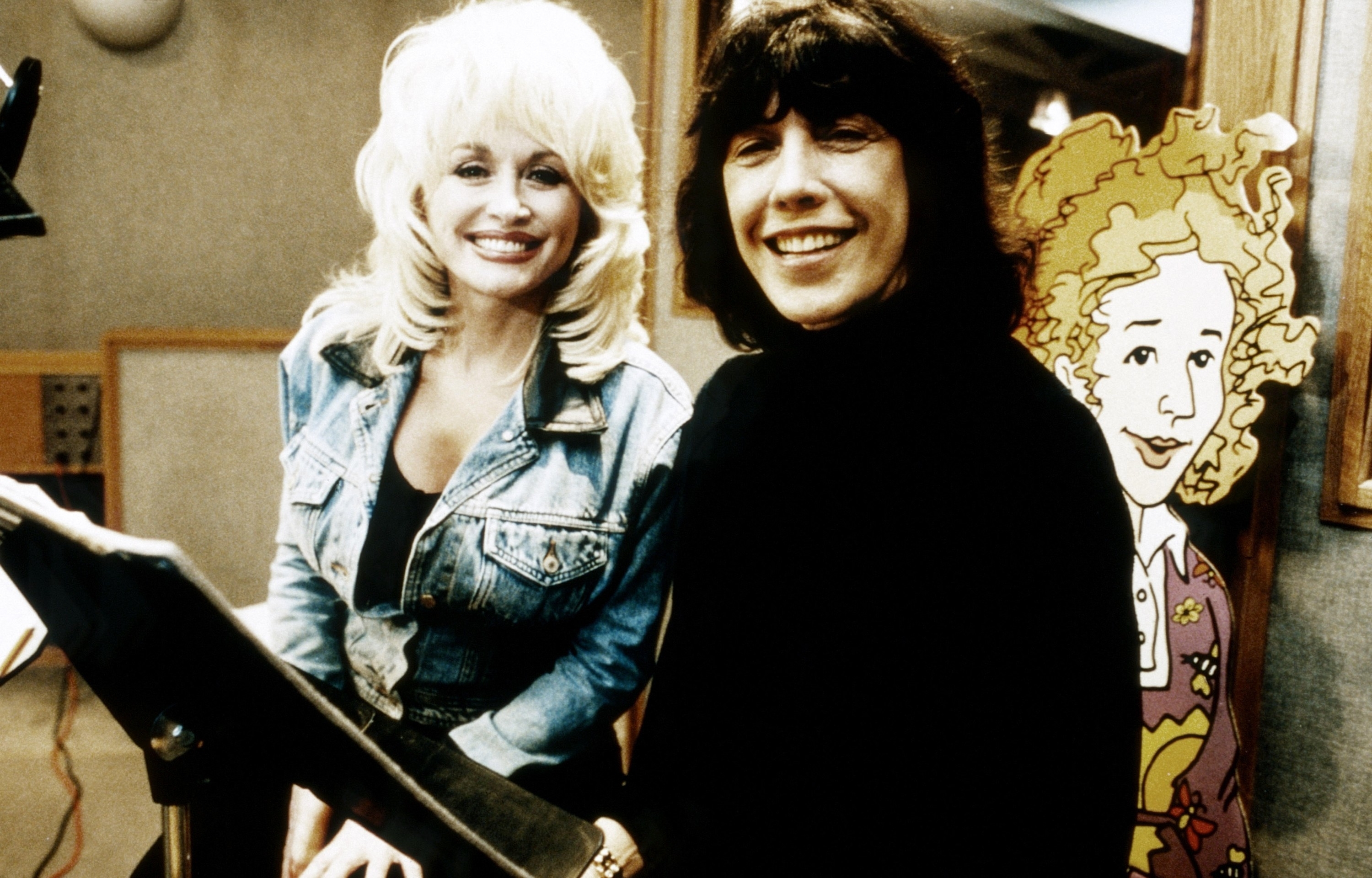 Dolly Parton, in a denim jacket, and Lily Tomlin, in a black outfit, are smiling in a recording studio with a cartoon character in the background