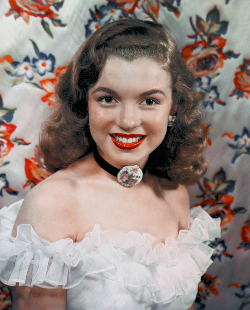 A young Marilyn Monroe smiles at the camera, dressed in an off-the-shoulder ruffled blouse, with a floral choker, against a floral backdrop