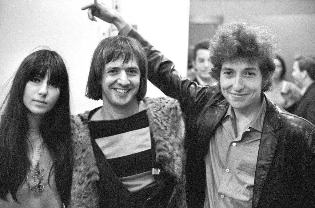 Cher, Sonny Bono, and Bob Dylan pose together at an event. Cher and Sonny wear casual outfits, and Bob Dylan sports a leather jacket