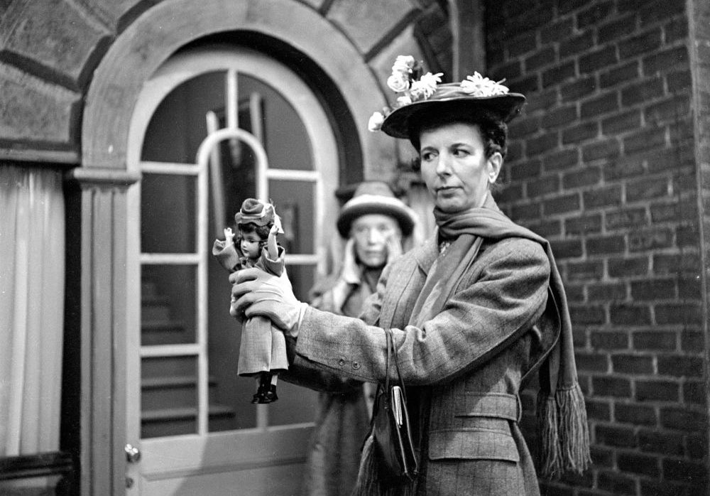 Two women in period attire stand outside a building. One holds a small puppet resembling the other woman
