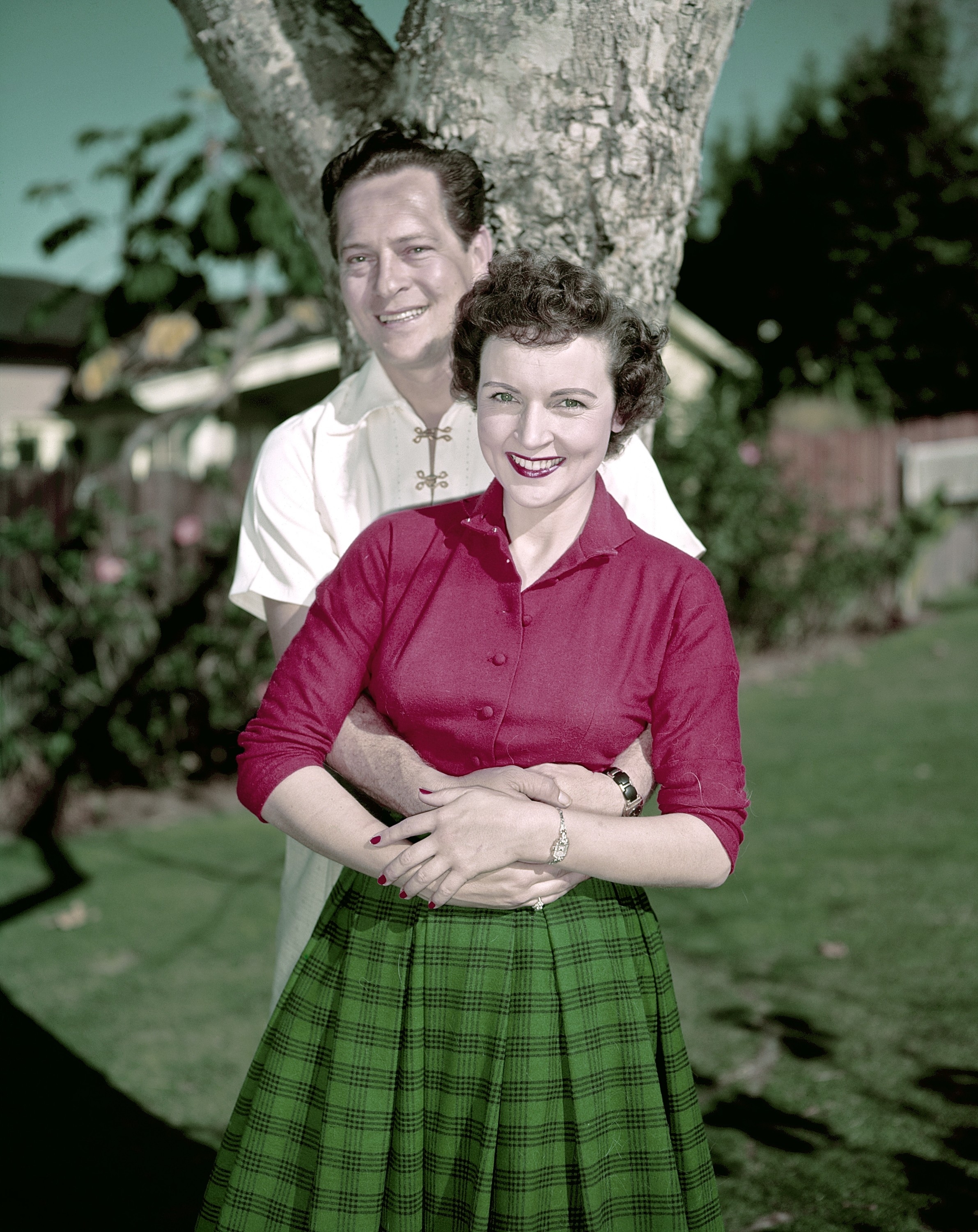 Betty White stands smiling in front of Allen Ludden, who is hugging her from behind near a tree in a garden
