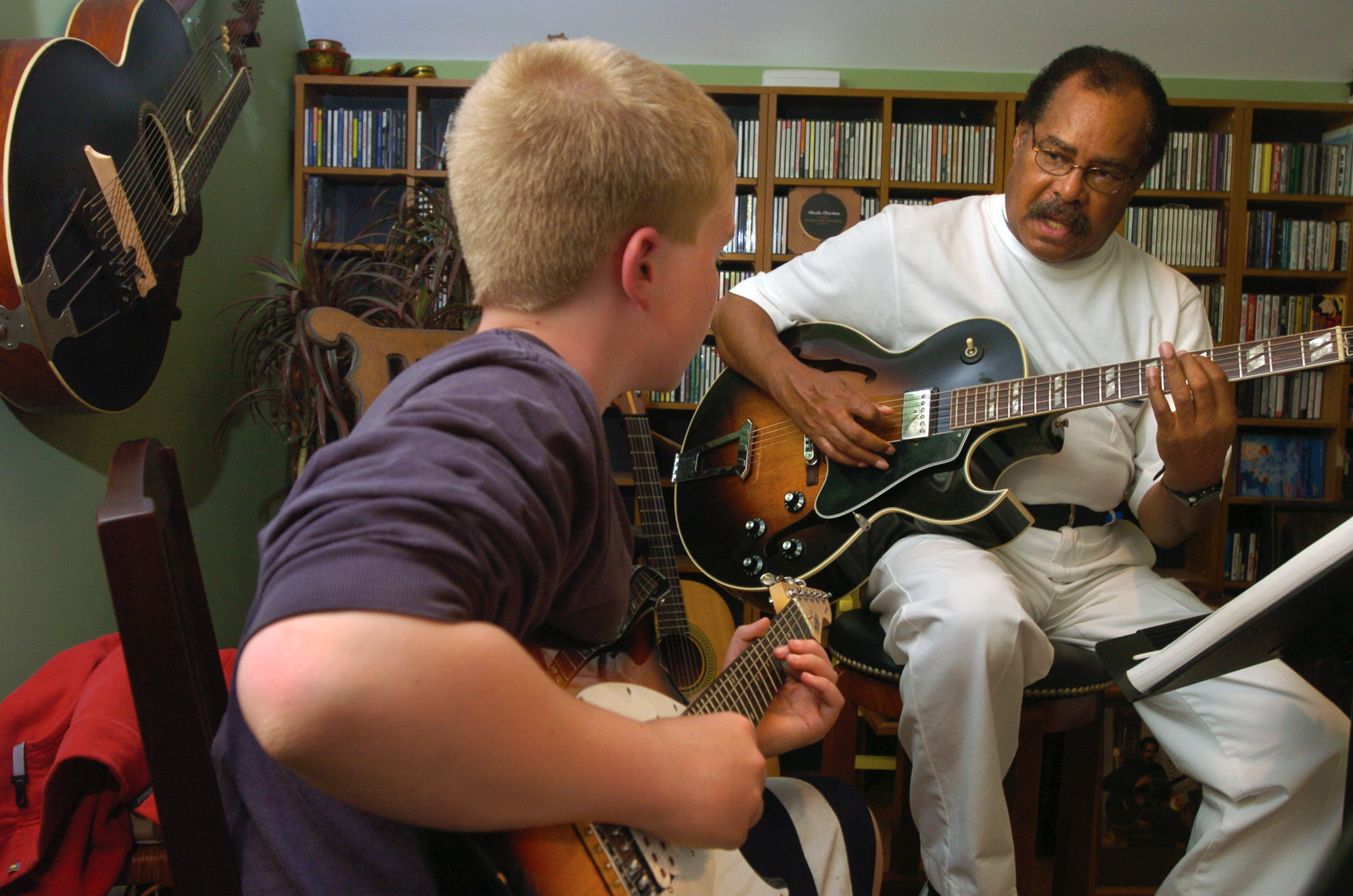 A man and a boy are playing guitars together in a room filled with CDs and guitars