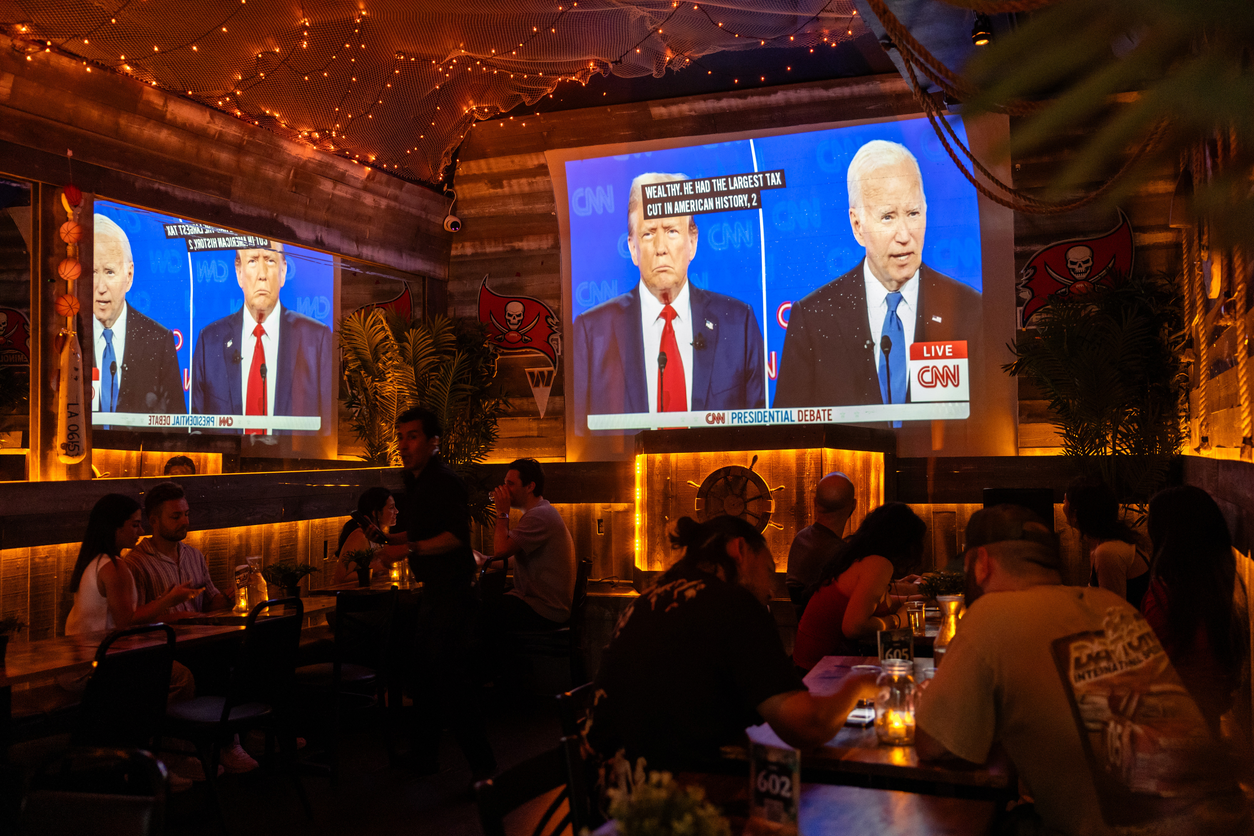 Television screens display Joe Biden and Donald Trump in a CNN presidential debate as people watch from tables in a dimly lit restaurant or bar