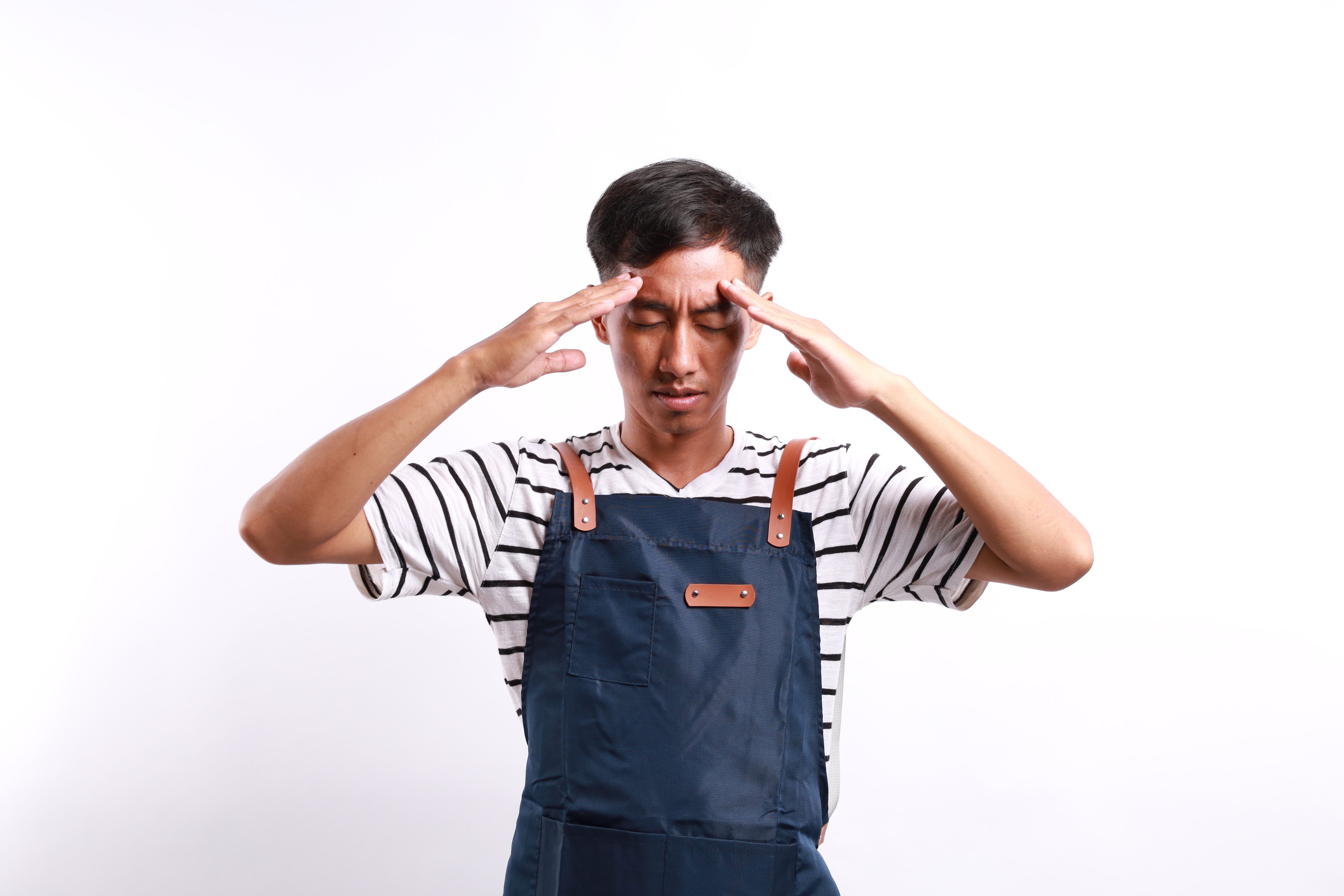 A person in a striped shirt and apron has their eyes closed, raising their hands with fingers touching their temples, appearing to be in deep thought or concentration