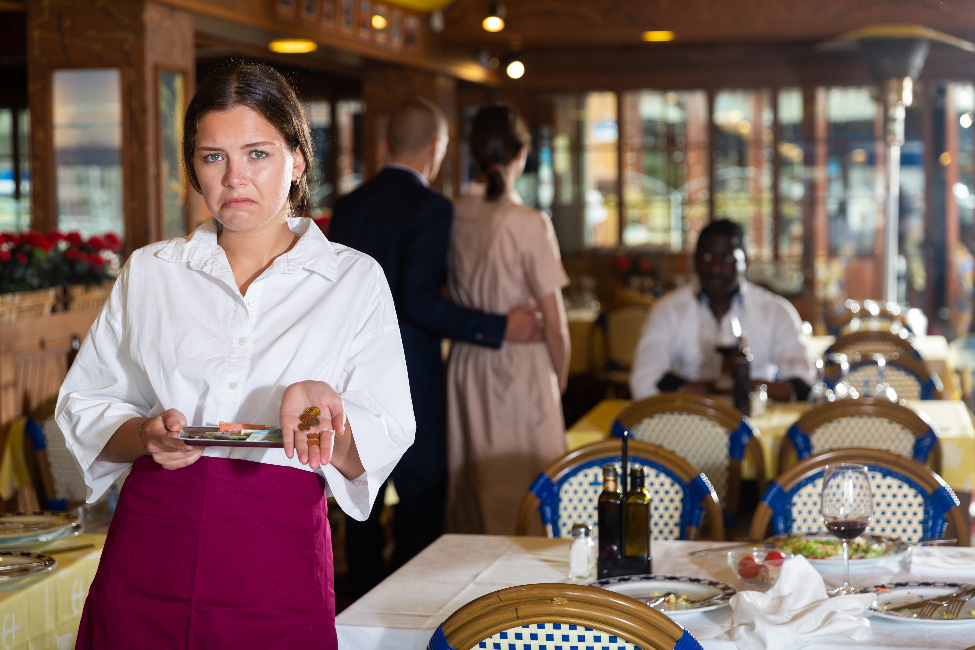 A restaurant waitress looks displeased while holding an empty tray. In the background, a couple embraces, and a man dines alone