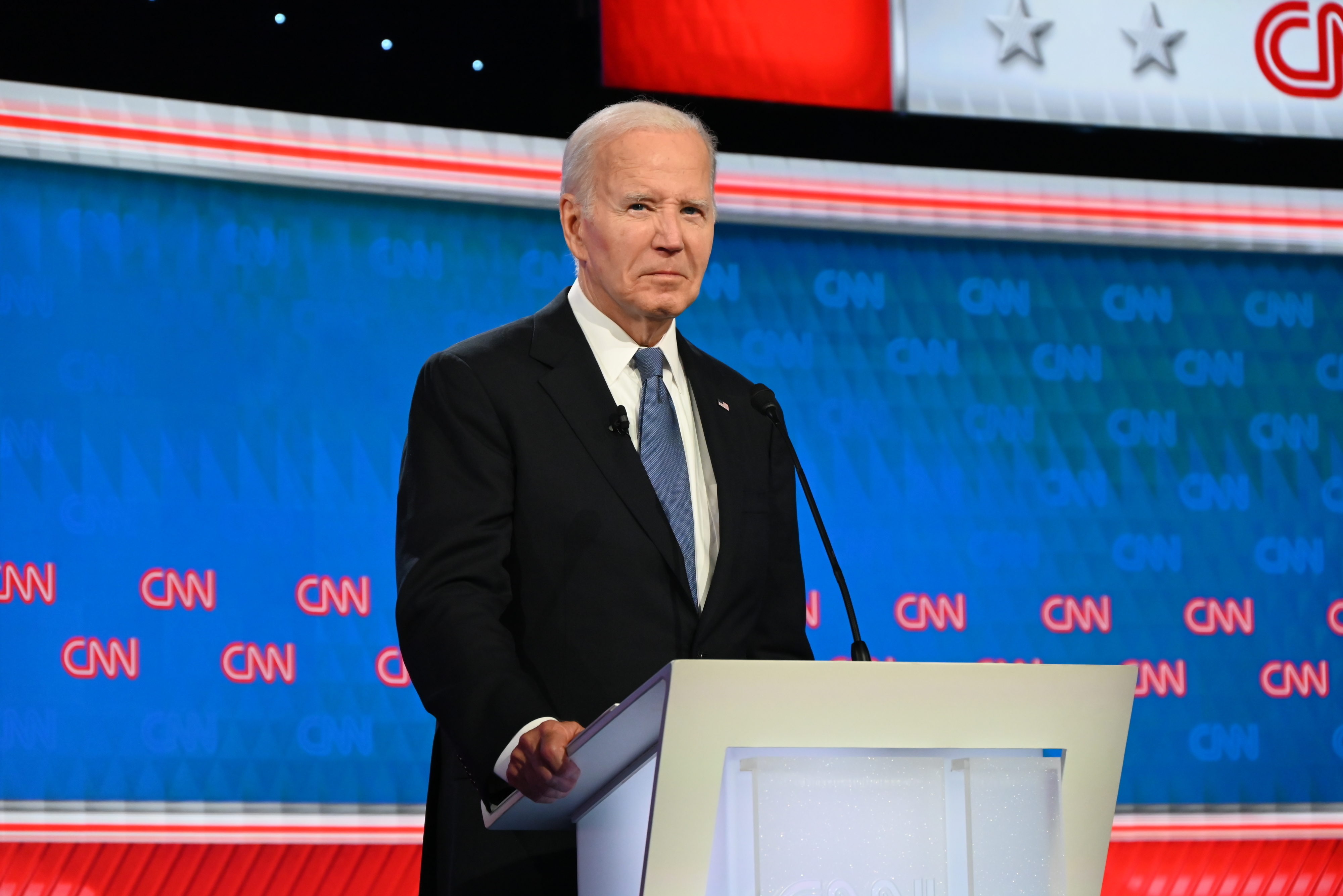 Joe Biden stands behind a podium at a CNN event, wearing a dark suit and blue tie, with CNN logos and stage lighting in the background