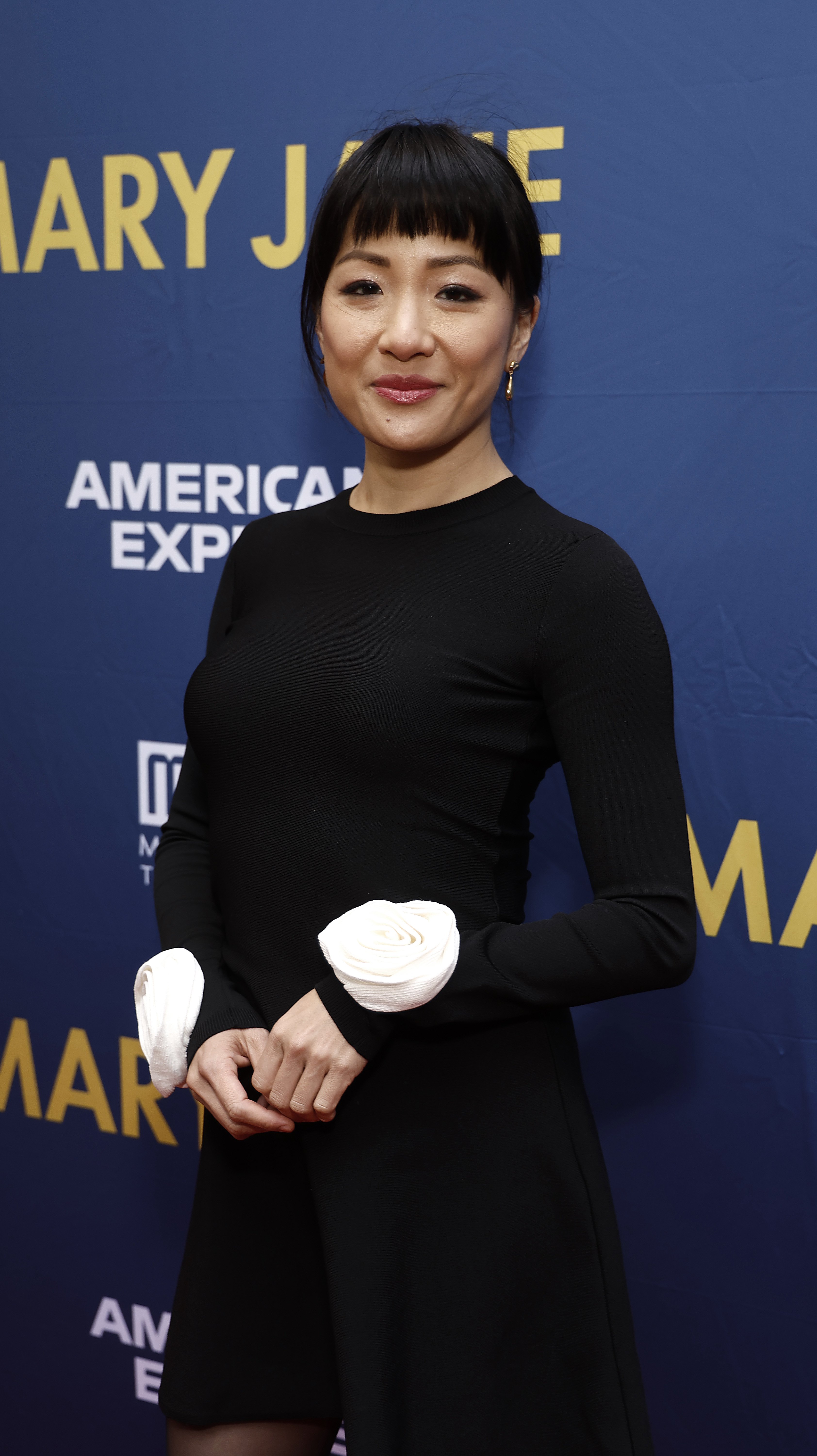 Constance Wu in a chic black dress with distinctive white accents on the sleeves, posing on a red carpet with &quot;Mary Jane&quot; and &quot;American Express&quot; logos in the background