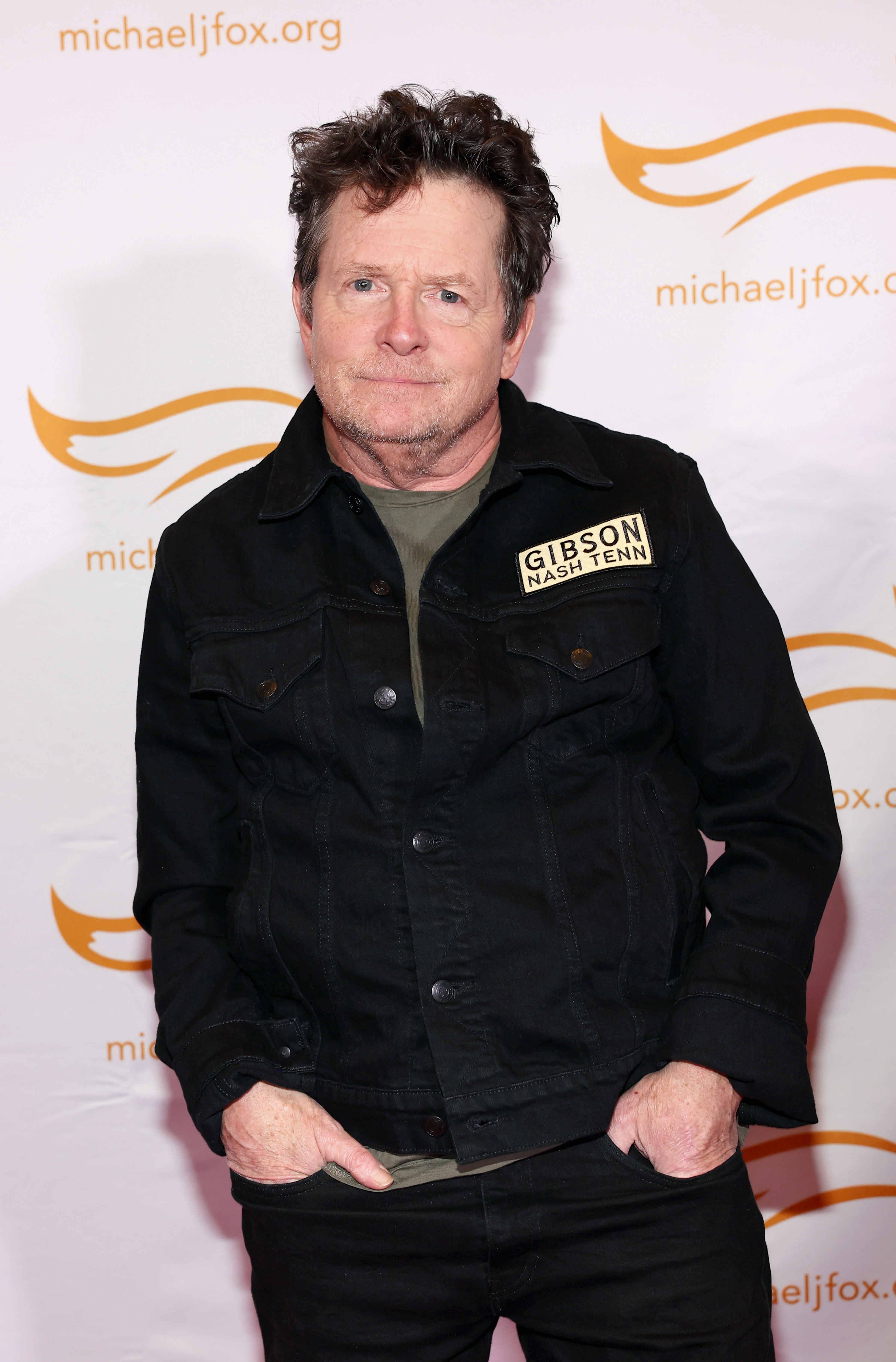 Michael J. Fox stands with hands in pockets, wearing a dark jacket with a &quot;Gibson Nash Tenn&quot; patch, in front of a backdrop with &quot;michaeljfox.org&quot; logos