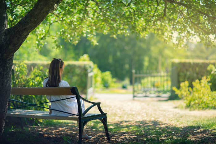 A person in white clothing with long brown hair sits on a bench near a tree in a tranquil garden. Sunlight filters through the leaves