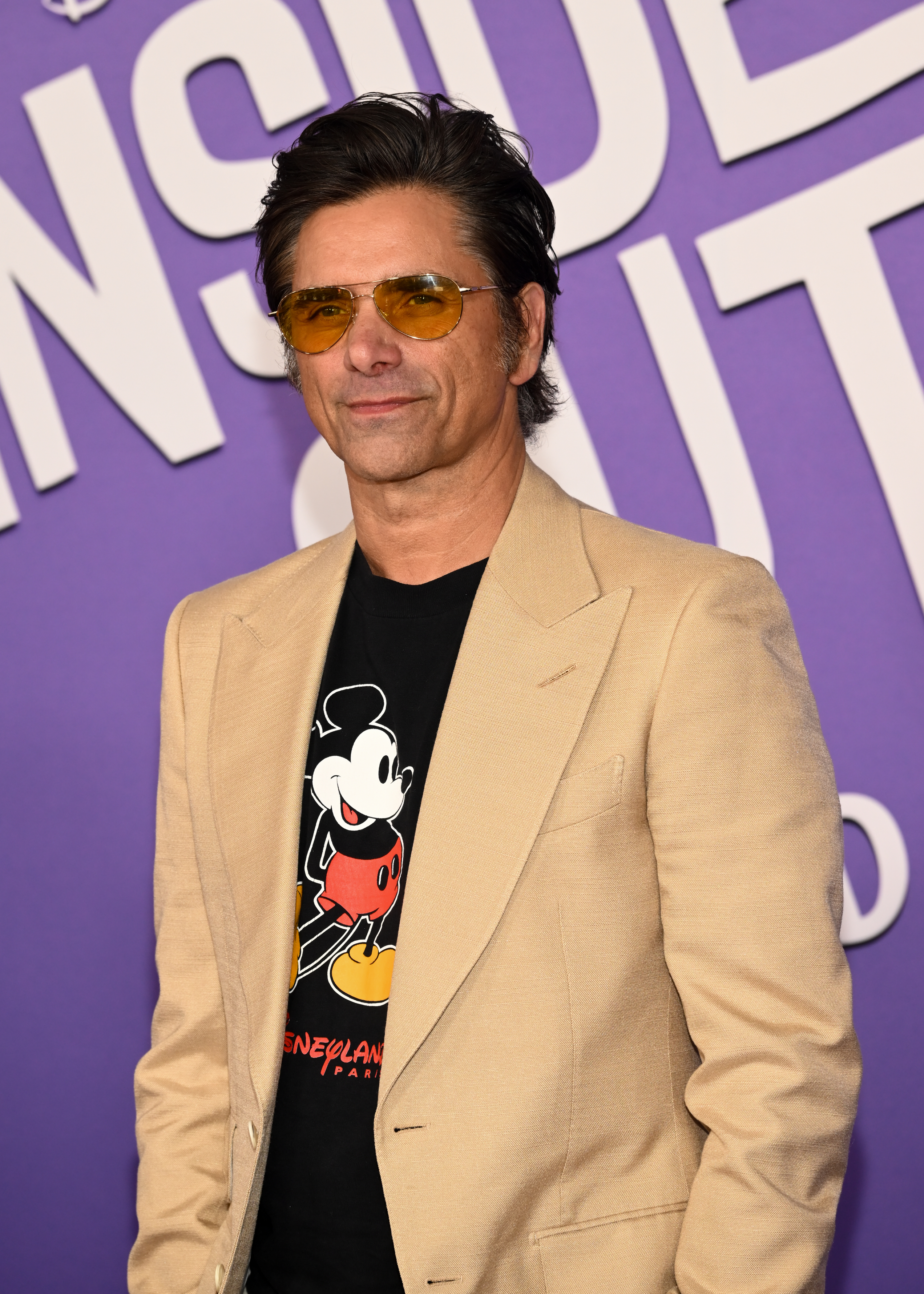 John Stamos wearing a Mickey Mouse T-shirt and beige blazer, attending a celebrity event in front of a large purple &quot;Inside Out&quot; sign