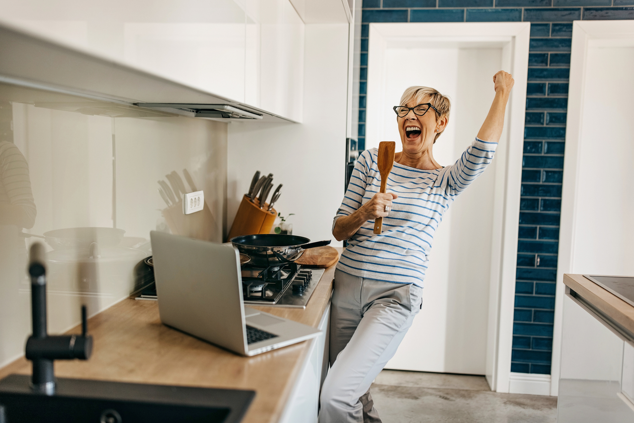 A person joyfully holds a wooden spoon in one hand and raises a fist in the other while standing in a modern kitchen beside a laptop on the counter