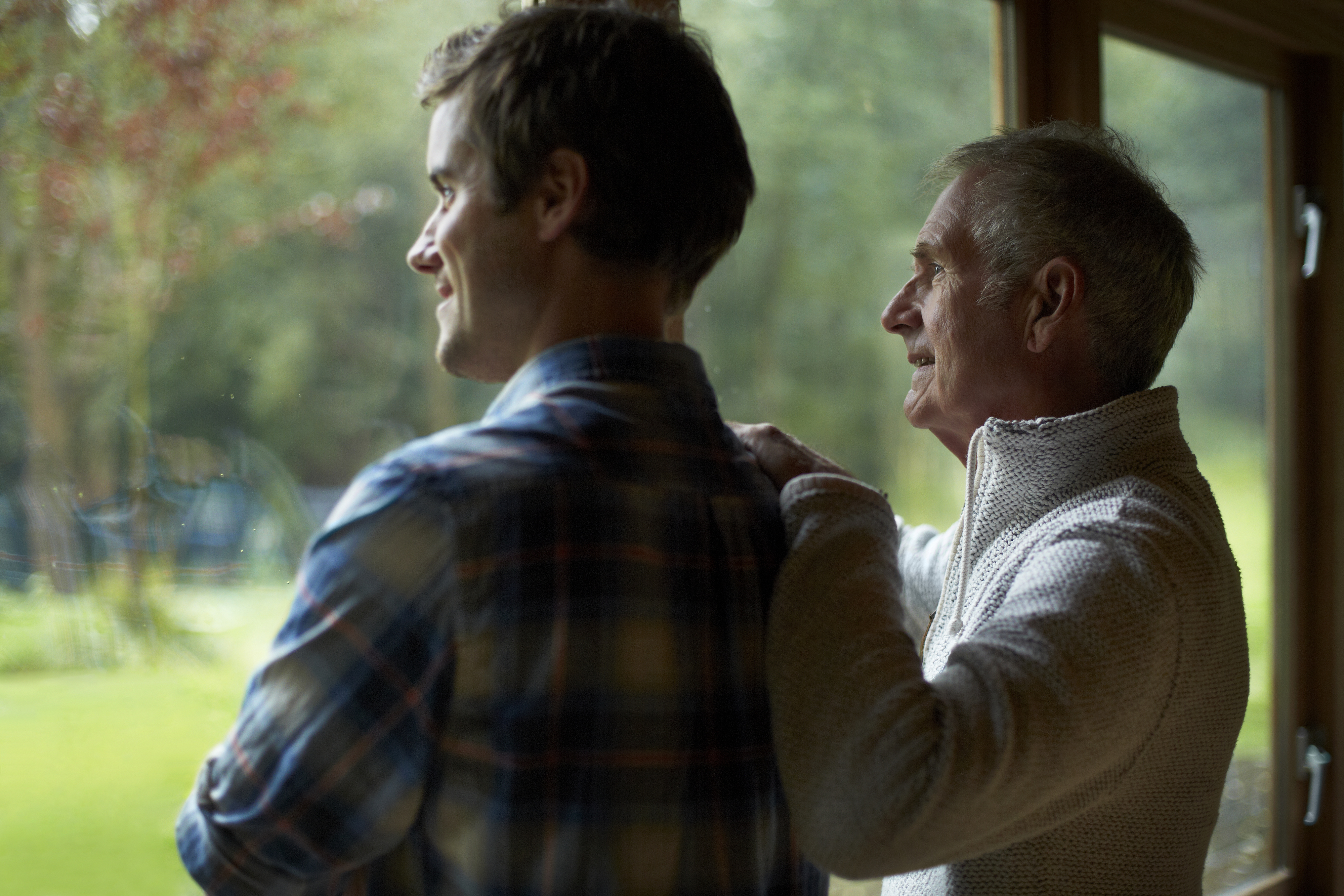 Two people stand together, one younger and one older, looking out a window with the older person resting their hand on the younger person&#x27;s shoulder. Names unknown