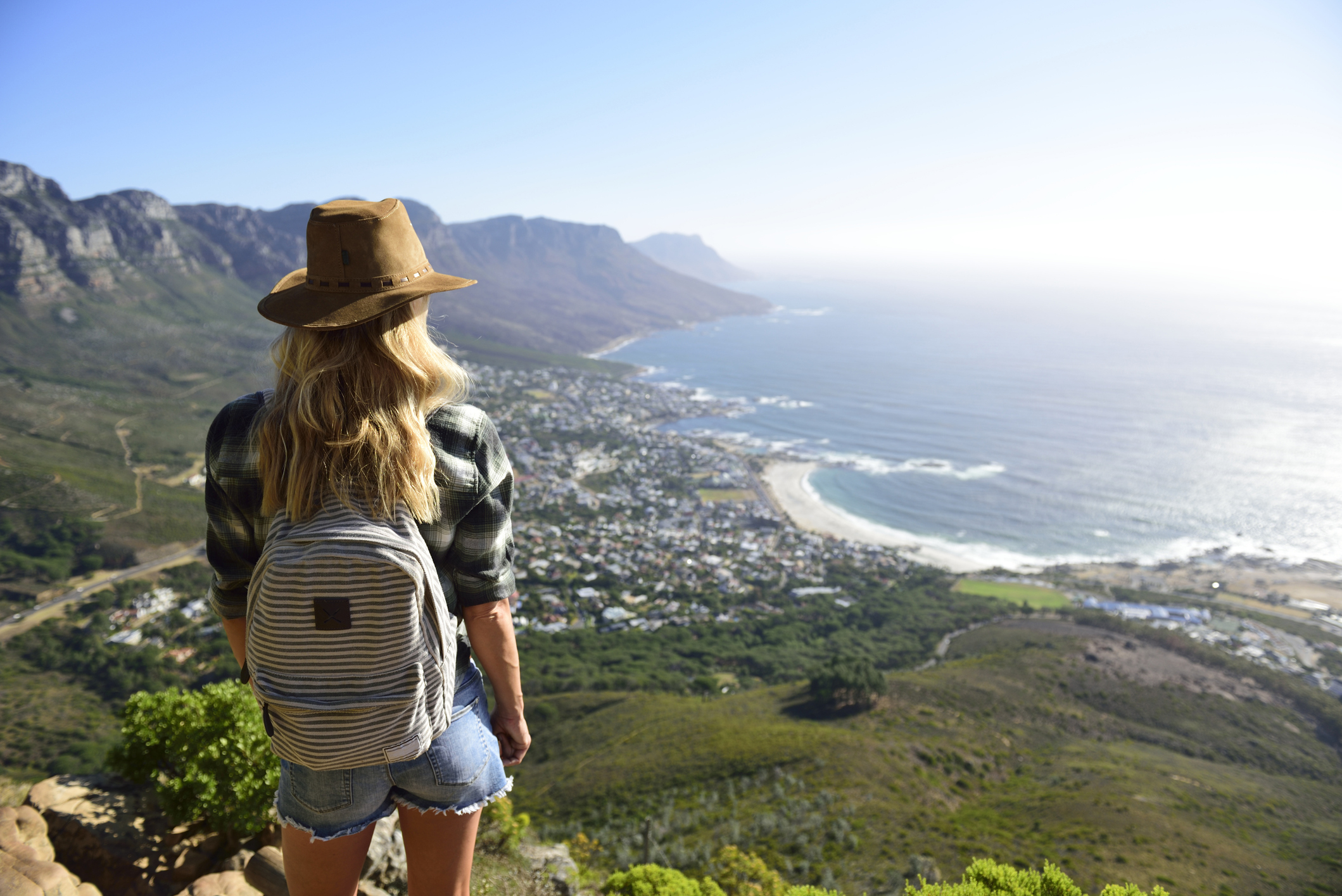 A woman with a hat and backpack overlooks a coastal town from a high vantage point, with a mountainous landscape in the background