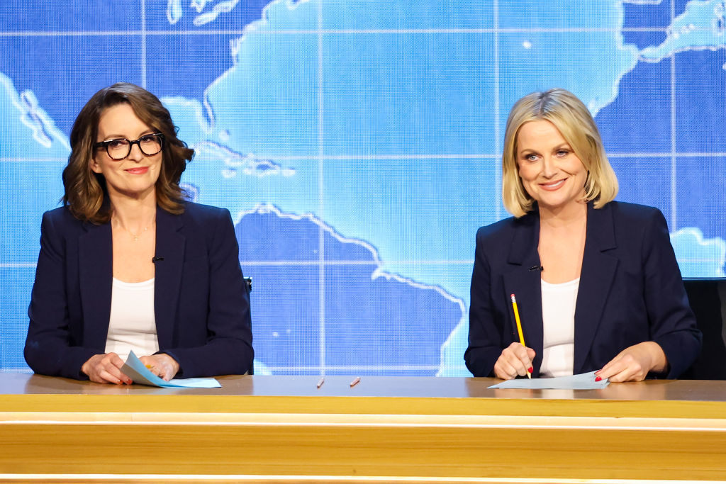 Tina Fey and Amy Poehler sit at a news desk with a world map background, both in white tops and dark blazers, holding papers and a pencil