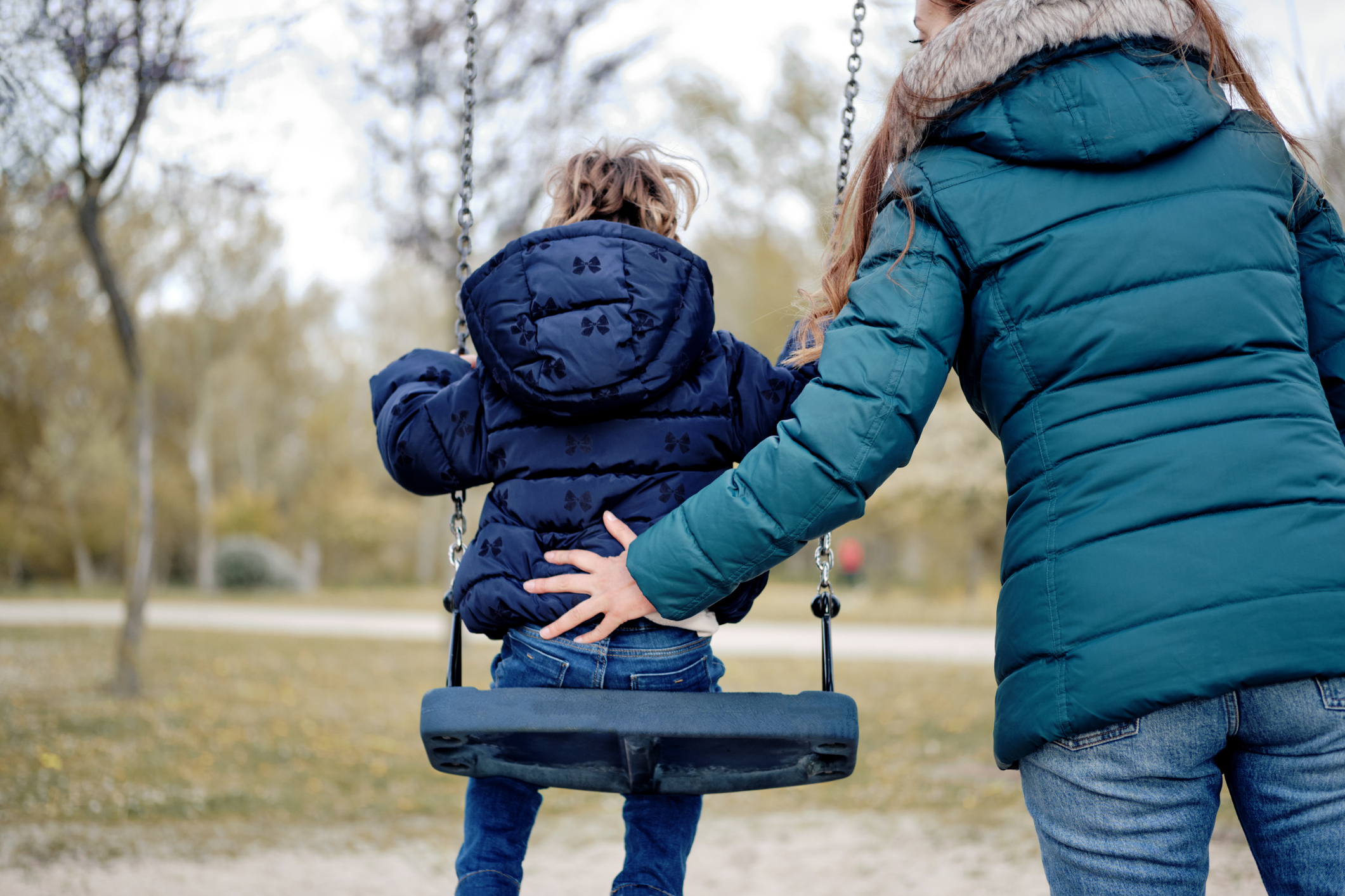 An adult pushes a child in a swing at a park. Both are wearing winter jackets