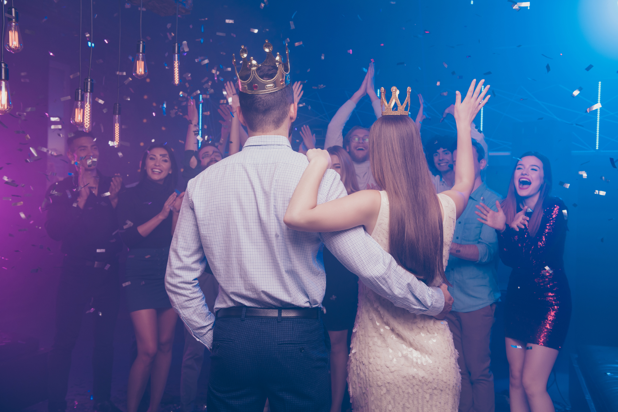 A man and woman wearing crowns stand arm in arm facing a cheering, confetti-filled crowd at a celebration event