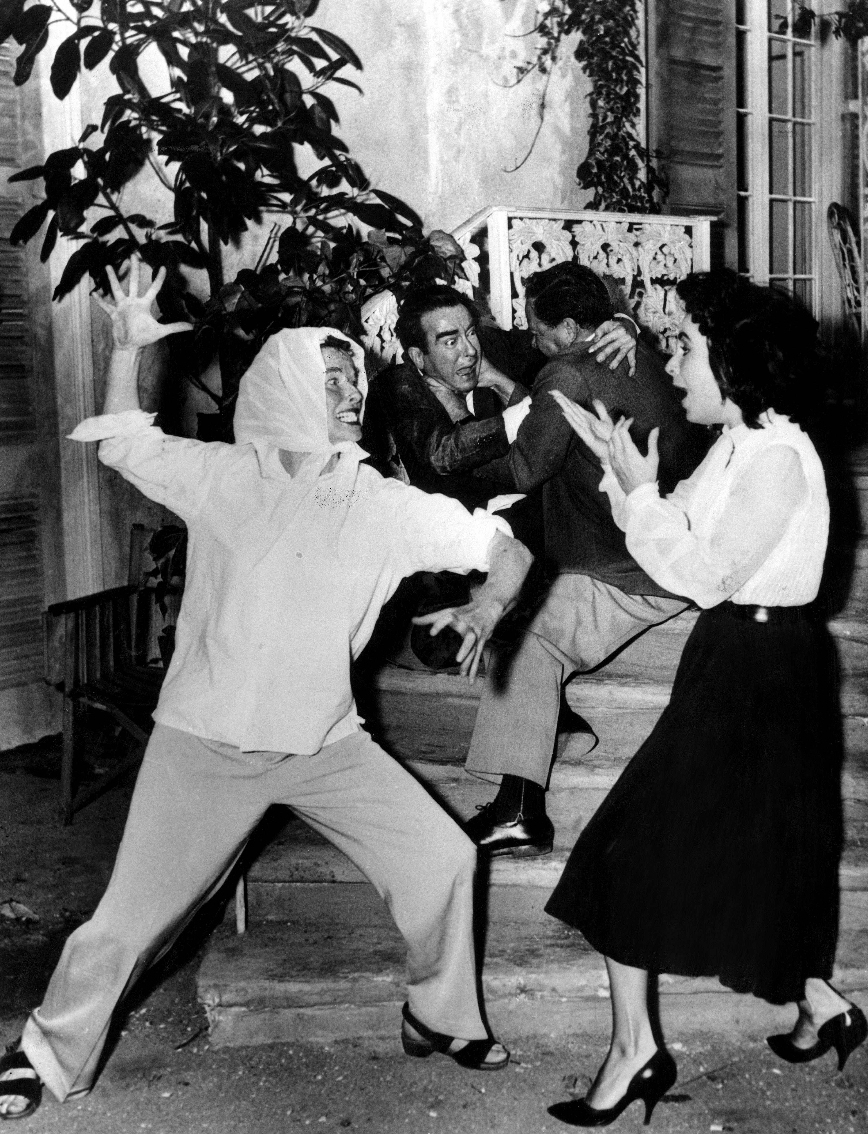 A behind-the-scenes, black-and-white shot of Elizabeth Taylor, Richard Burton, and Rex Harrison playfully acting during a film&#x27;s production