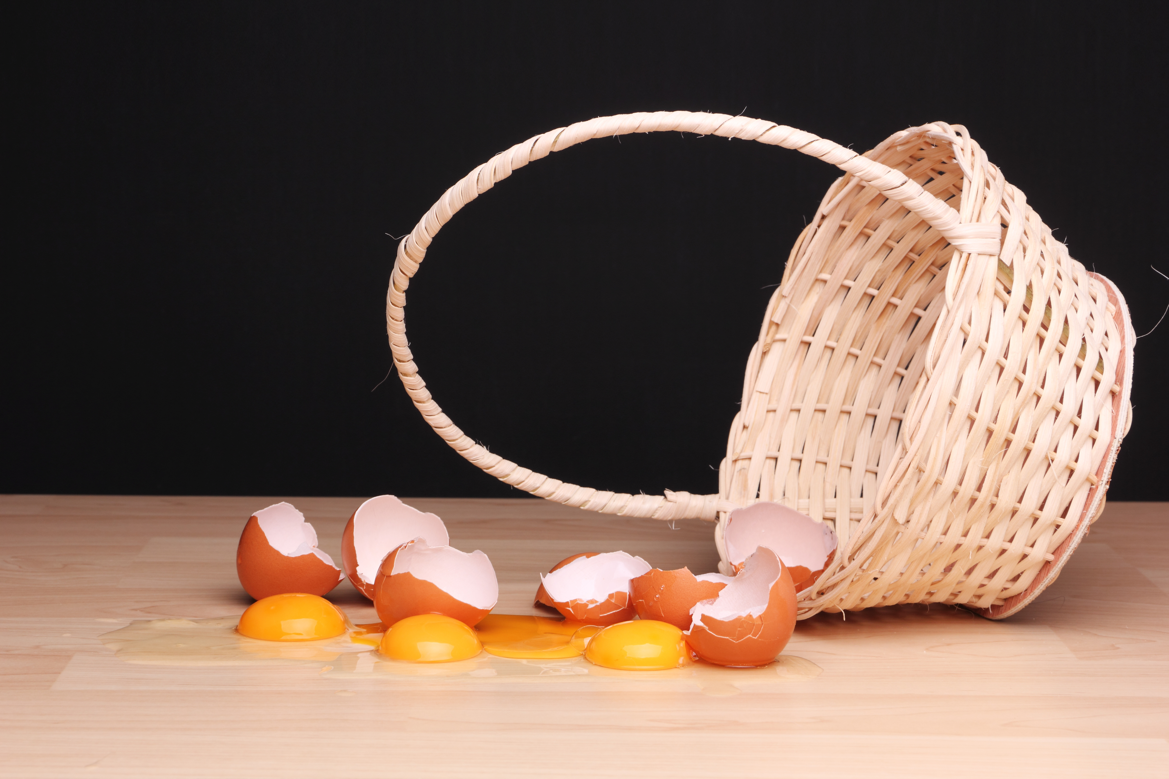 Broken eggs and eggshells are spilled from an overturned wicker basket on a wooden surface