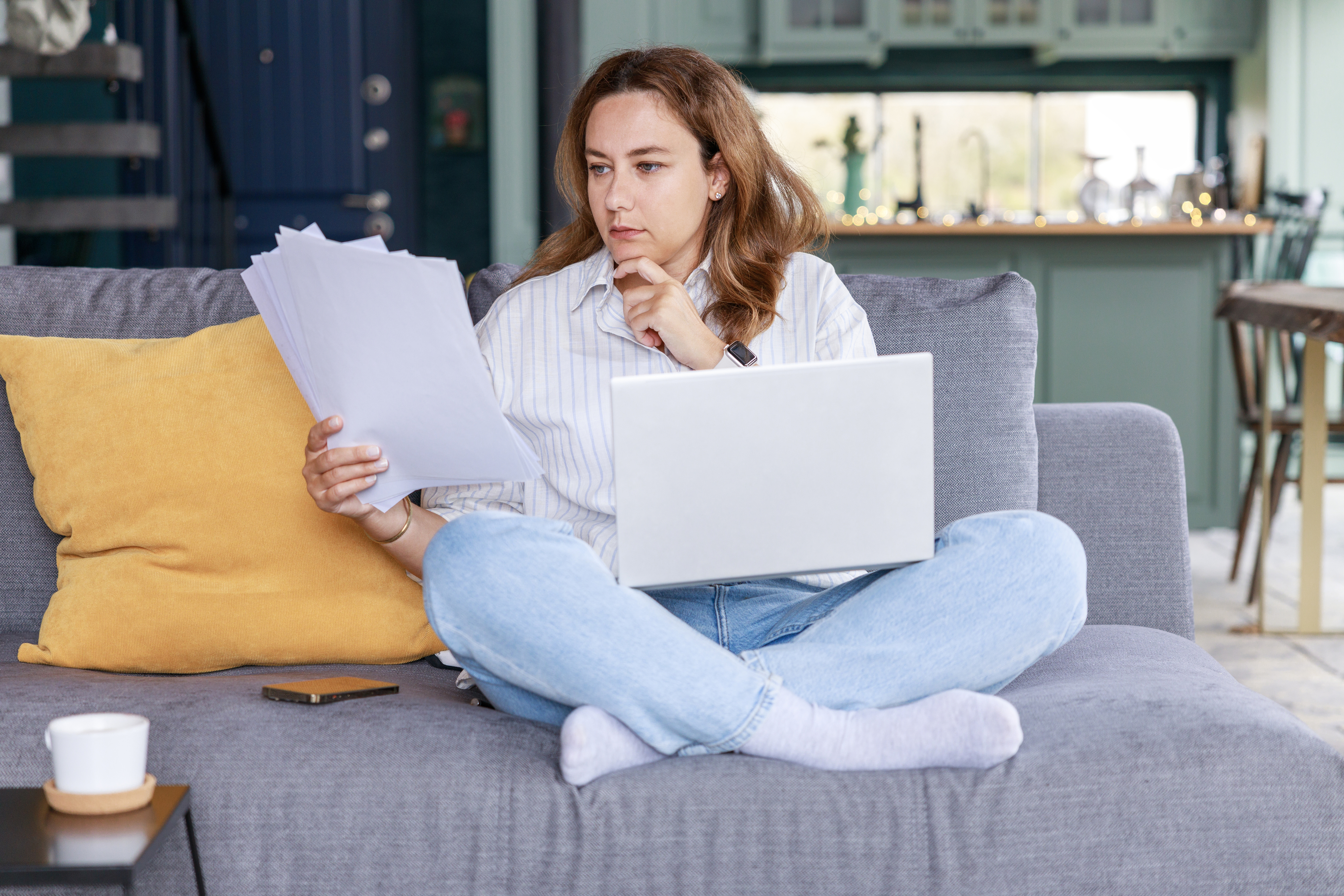 A person sits on a couch with a laptop on their lap, holding and examining a stack of papers with a thoughtful expression