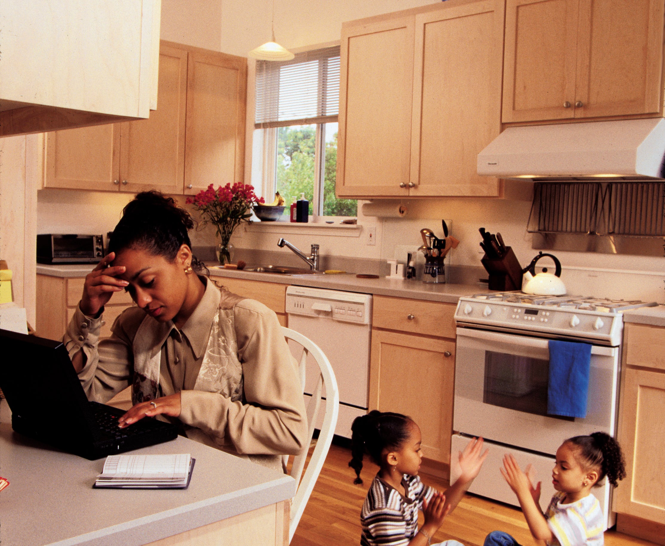 A woman sits at a kitchen table working on a laptop while two children play on the floor nearby