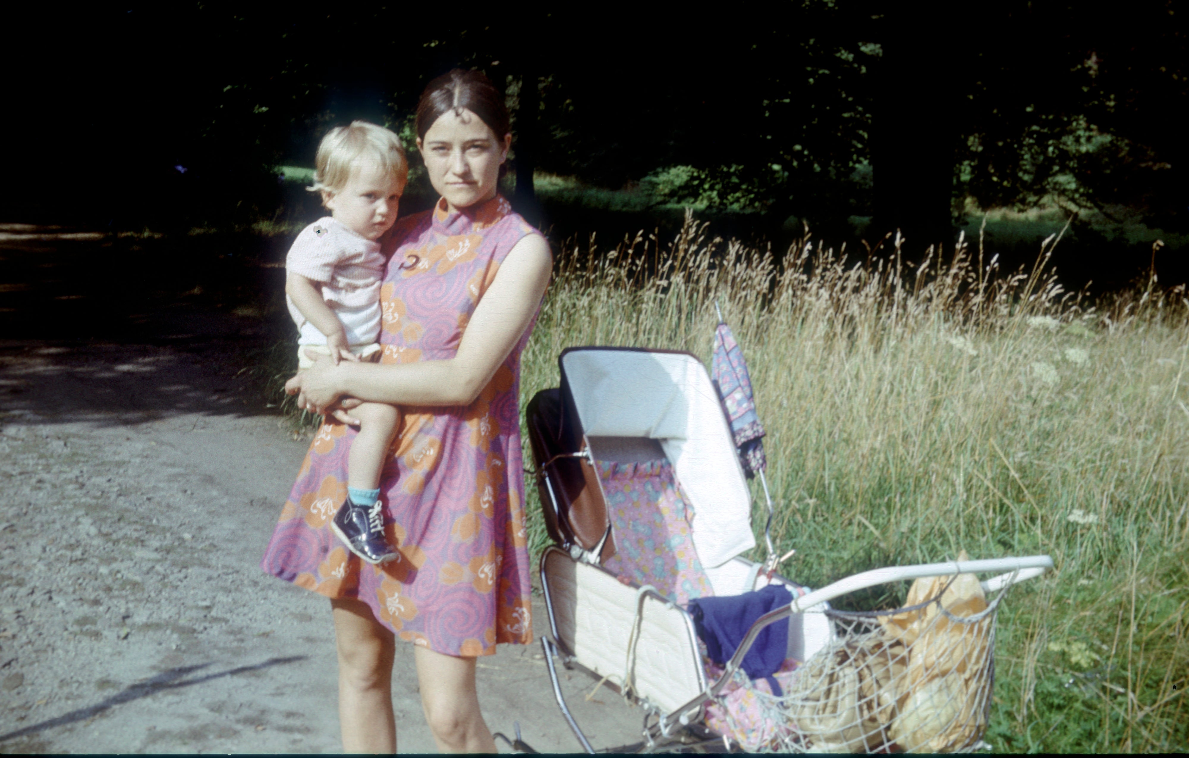 A woman in a patterned dress holds a young child next to a pram on a rural path