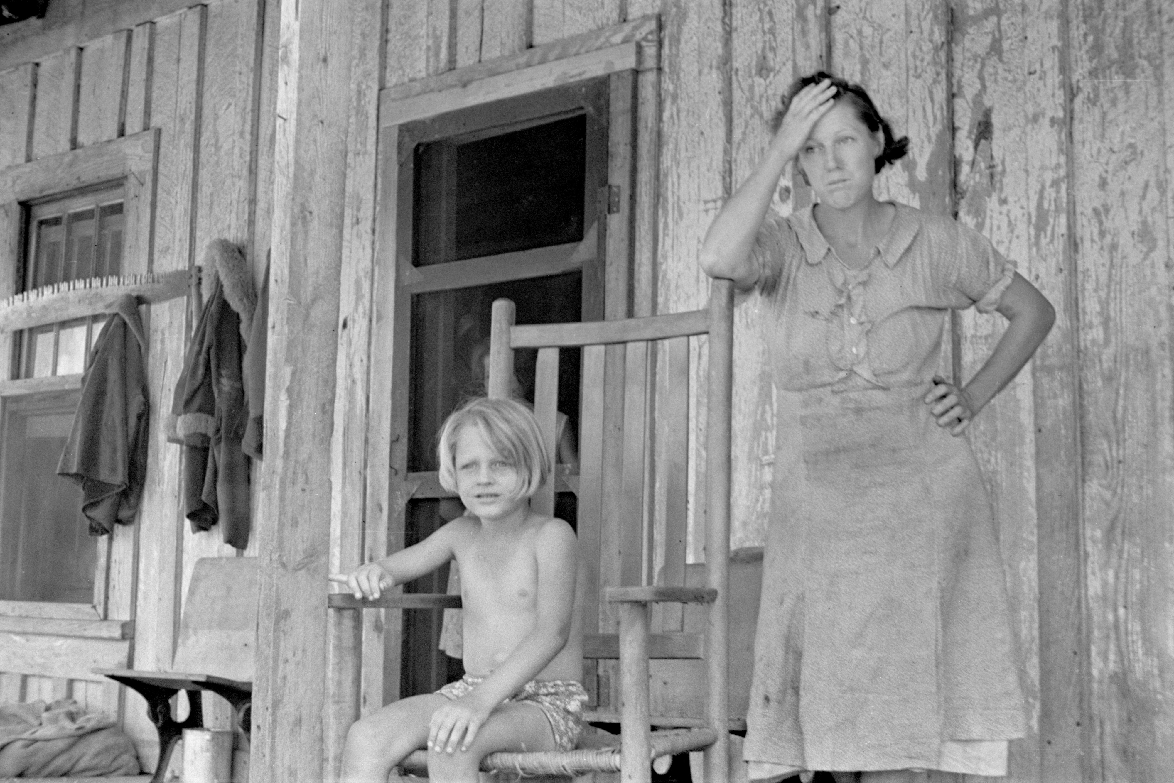 A woman stands with her hand on her forehead next to a shirtless child sitting on a wooden rocking chair on a rustic wooden porch