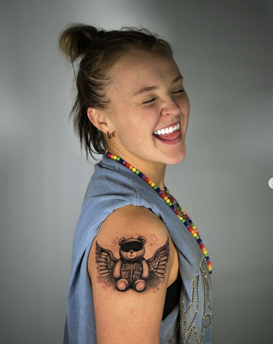JoJo Siwa smiling with eyes closed, showing a tattoo of a teddy bear with wings on her upper arm. She is wearing a sleeveless top and colorful bead necklace