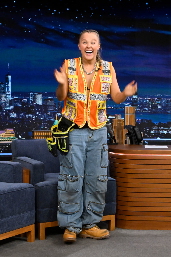 JoJo Siwa stands on a talk show stage, smiling and wearing a colorful vest, cargo pants, boots, and a toolbelt as she gestures energetically