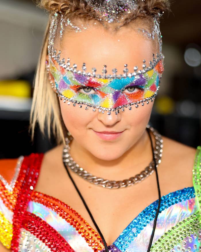 JoJo Siwa wearing a colorful, bejeweled outfit and an elaborate, multicolored glitter mask at an event