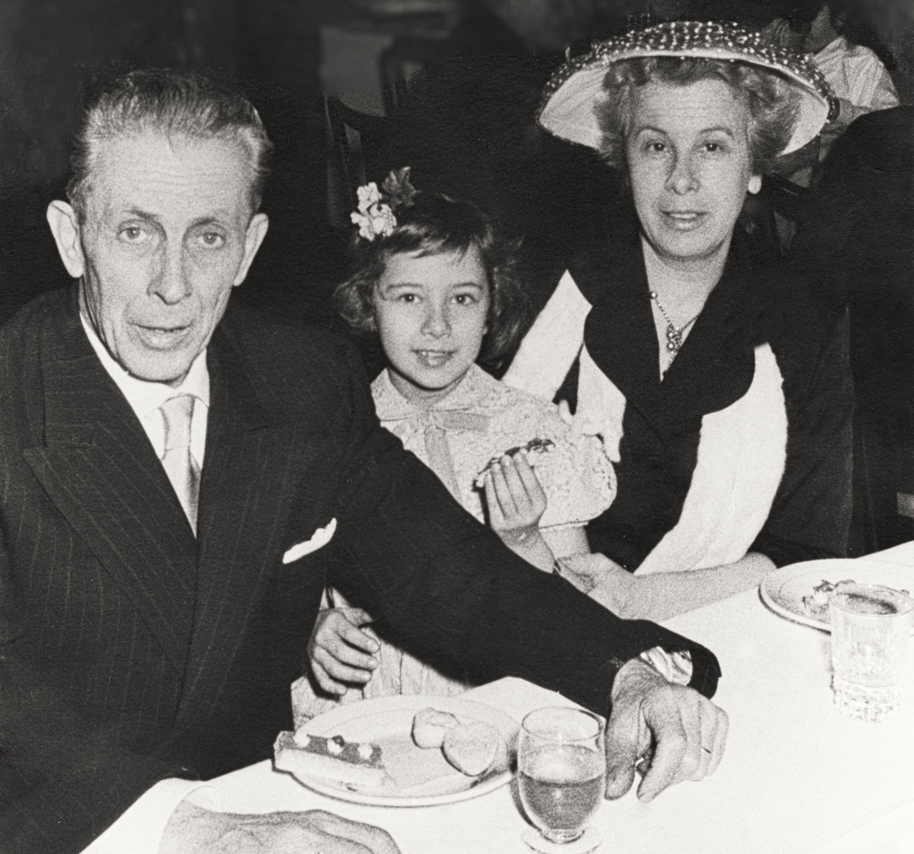 An adult male, a young girl, and an adult female are seated at a dining table, smiling at the camera. The woman is wearing a hat. Names of persons are unknown