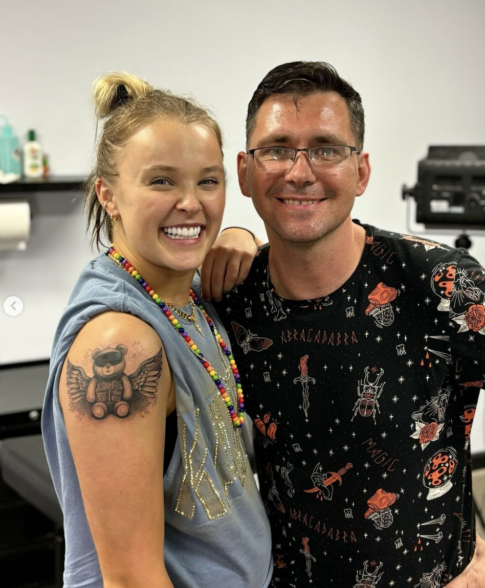 JoJo Siwa smiles, showing a new teddy bear tattoo on her arm, standing next to a man in an astrology-themed t-shirt. Both are posing indoors