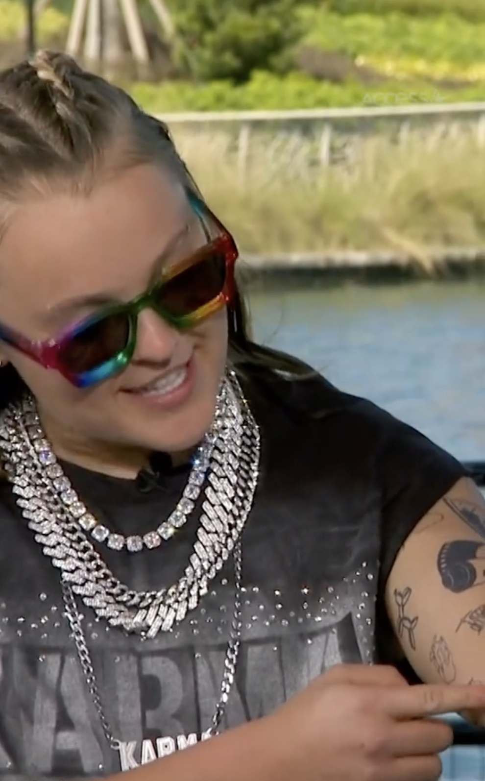 A person wearing large sunglasses, a T-shirt, and multiple layered necklaces shows off tattoos on their arm