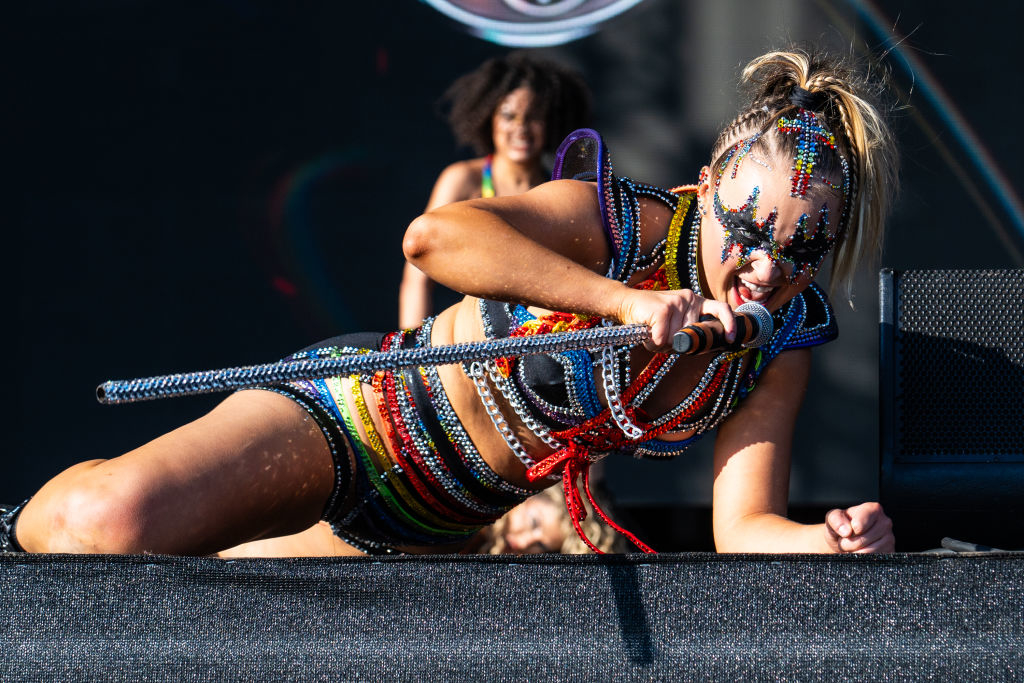 A performer in a beaded outfit and face decoration kneels while singing into a microphone on stage. Another person is visible in the background