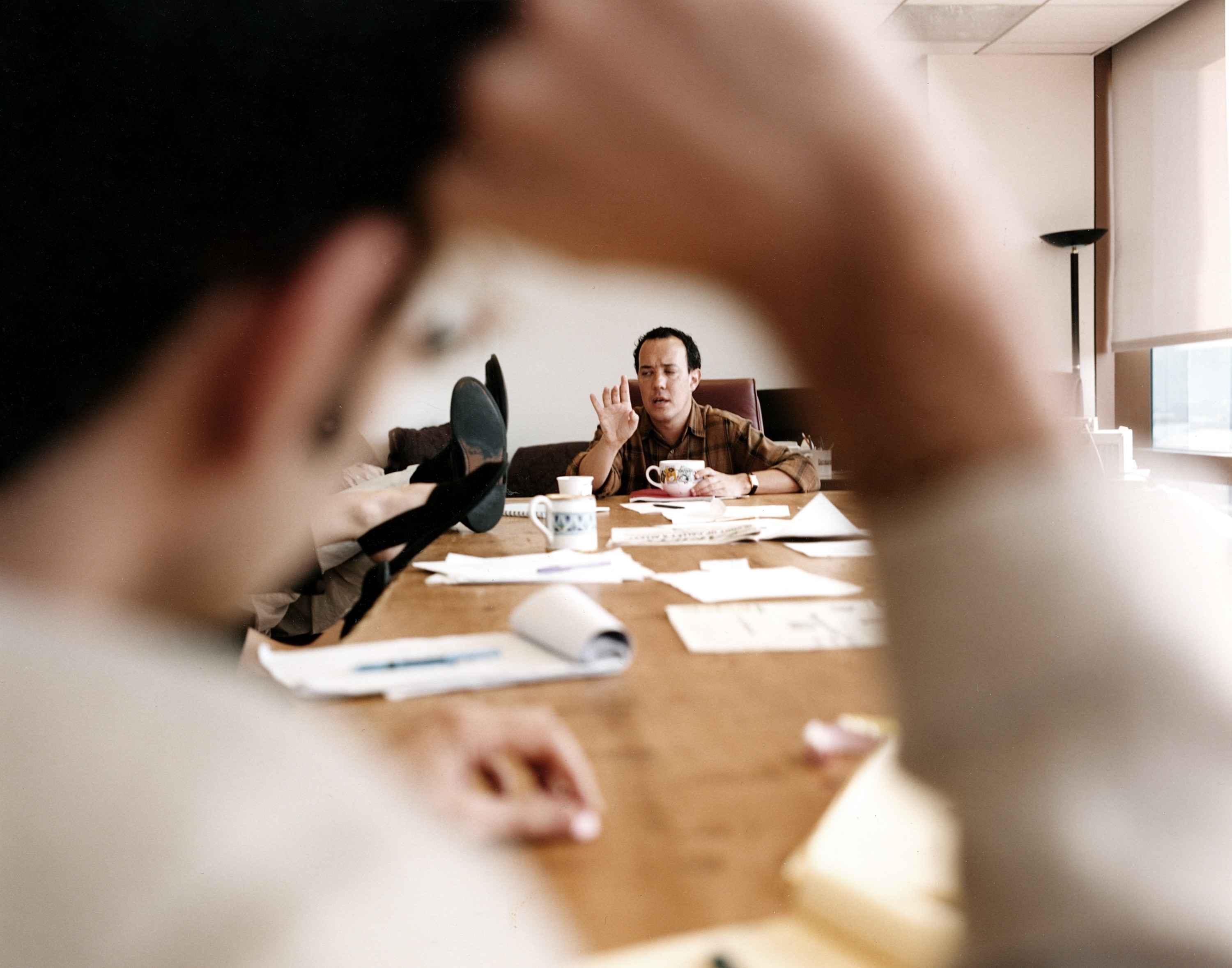 A man sits at a large, cluttered meeting table, engaging in a discussion. Two other people&#x27;s blurred figures are visible in the foreground
