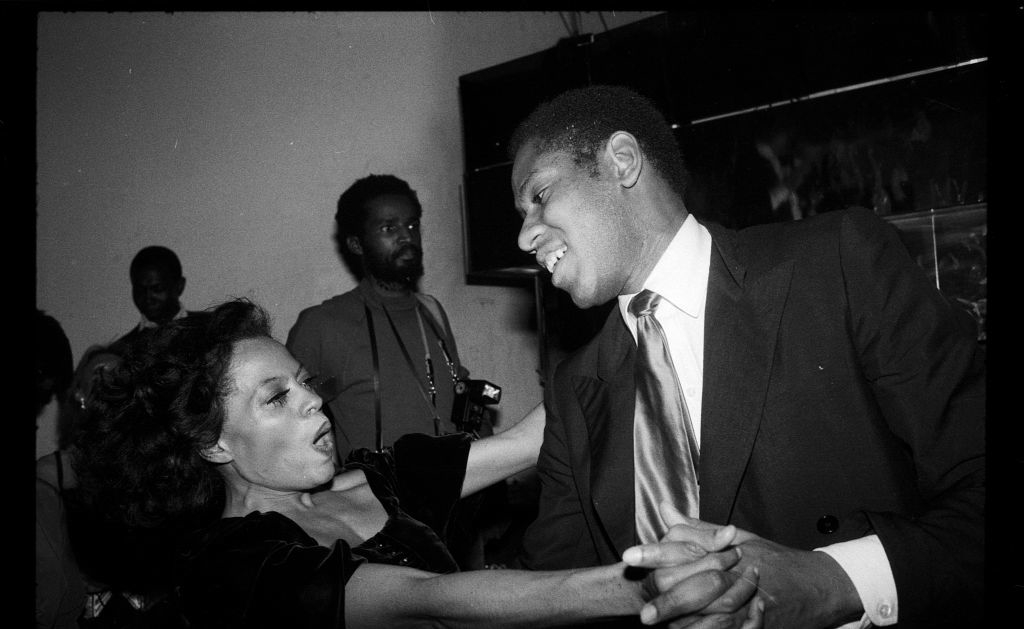 Diana Ross and André Leon Talley dancing enthusiastically at a social gathering. Diana is in a velvet dress, and André is wearing a suit and tie