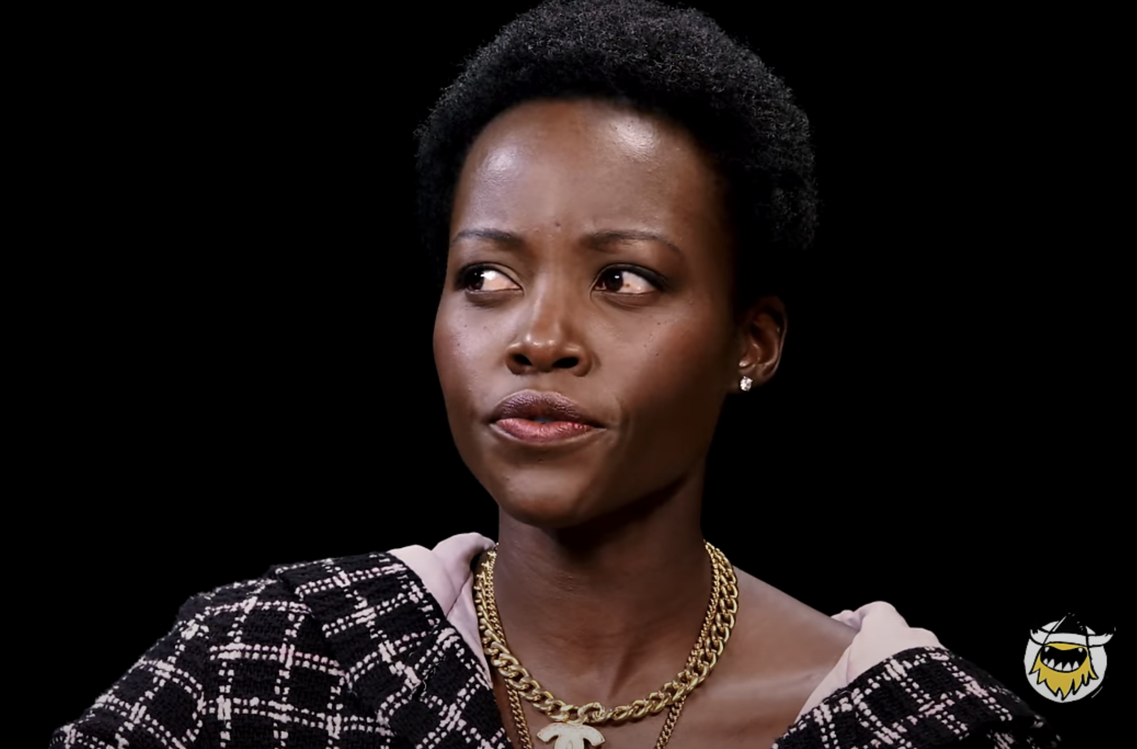 Lupita Nyong&#x27;o wearing a Chanel outfit and a gold Chanel necklace appears on the show &quot;Hot Ones&quot; with a Skeptical expression