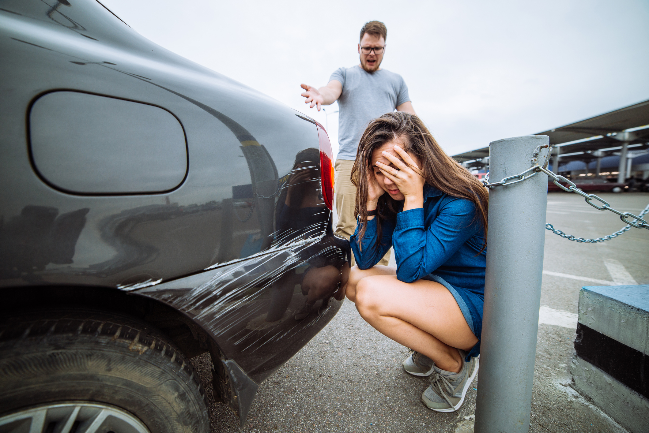 A distressed woman crouches by a scratched rear bumper of a car, covering her face. A man stands behind her, pointing at the damage with a frustrated expression