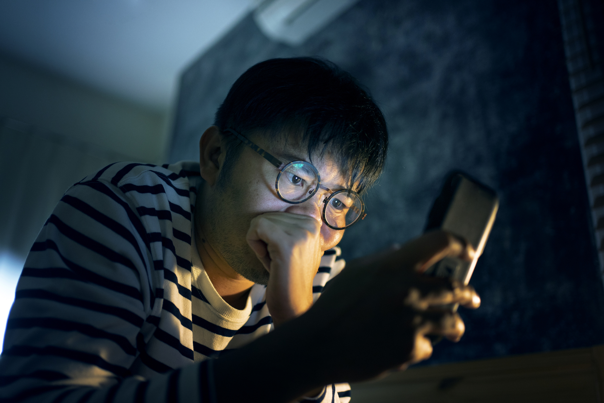 A person wearing glasses and a striped shirt looks intently at a smartphone screen, resting their chin on their hand