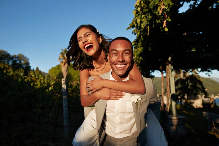 A woman joyfully rides piggyback on a man in a vineyard. Both are smiling widely, with the man wearing a light-colored suit and the woman in a sleeveless dress