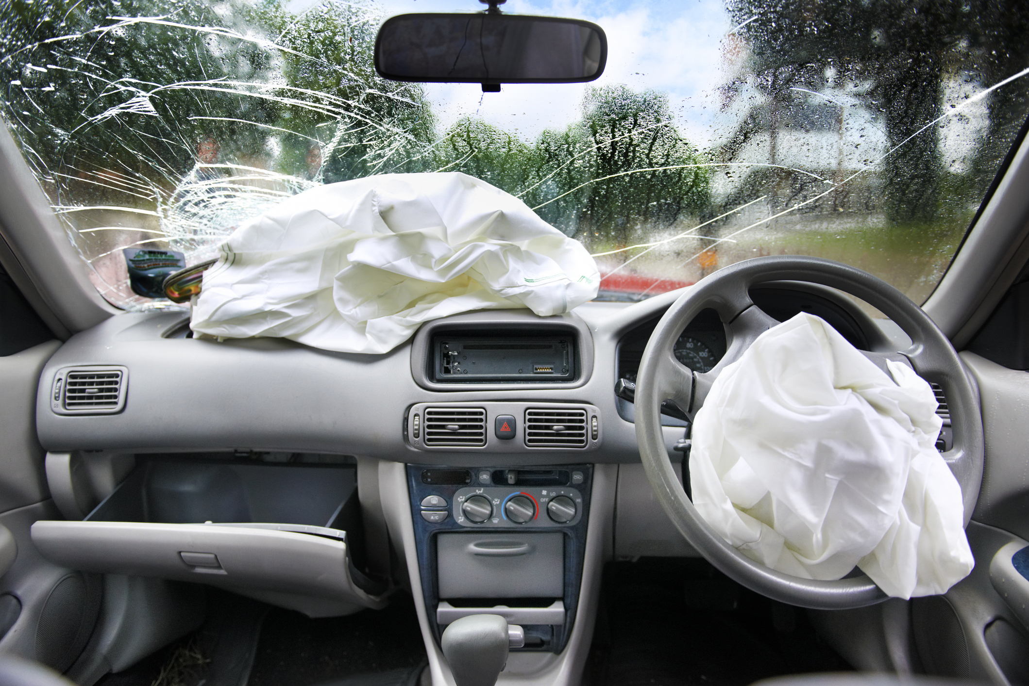 A car&#x27;s interior after an accident, showing deployed airbags and a shattered windshield. The vehicle&#x27;s dashboard, steering wheel, and broken glass are visible