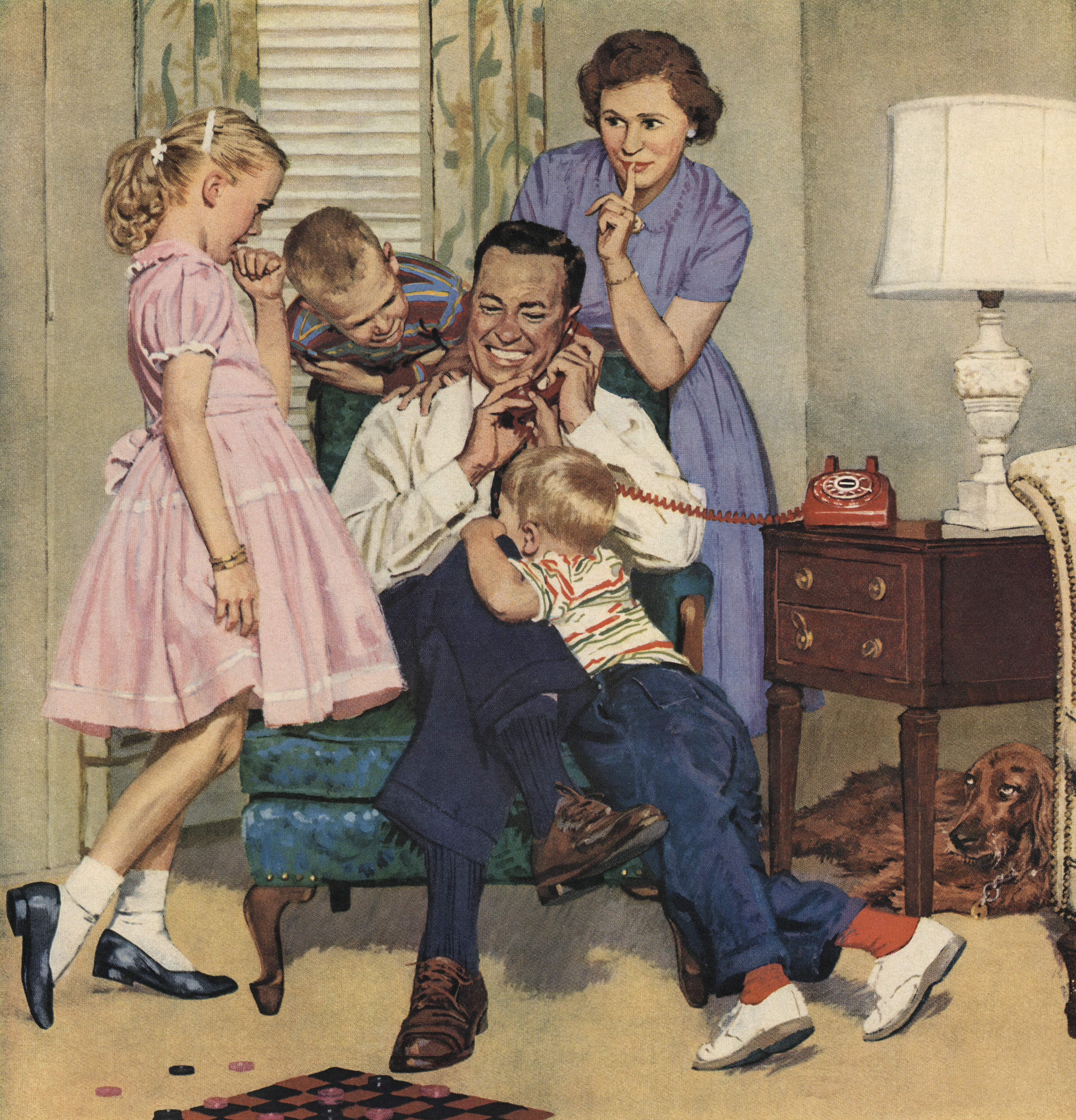 Father sits in a chair talking on the phone while his wife and three children playfully interact with him. A dog lies on the floor near a lamp and side table