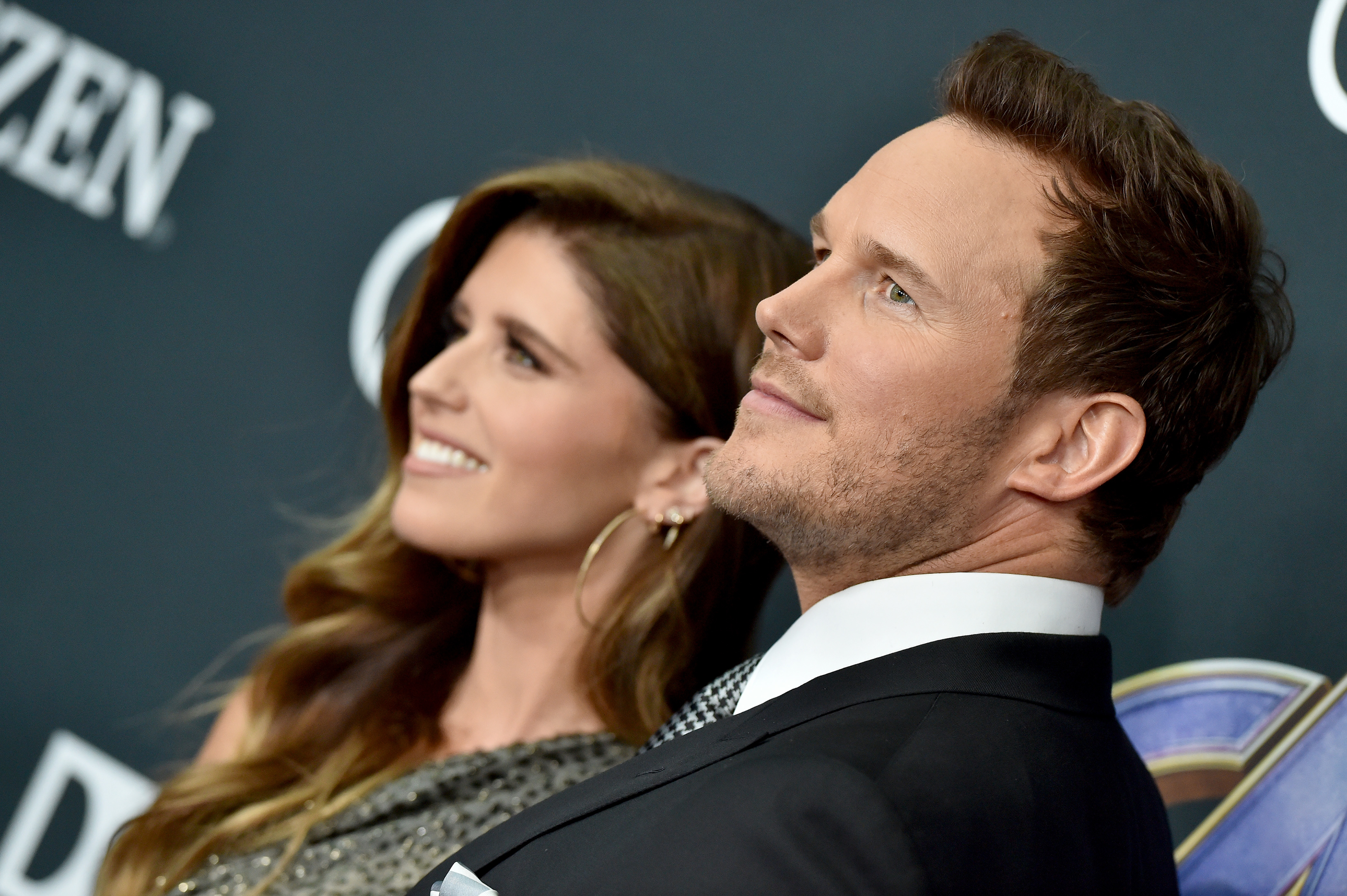 Chris Pratt and Katherine Schwarzenegger smiling and looking into the distance at a formal event. Chris is in a suit; Katherine is in a one-shoulder dress