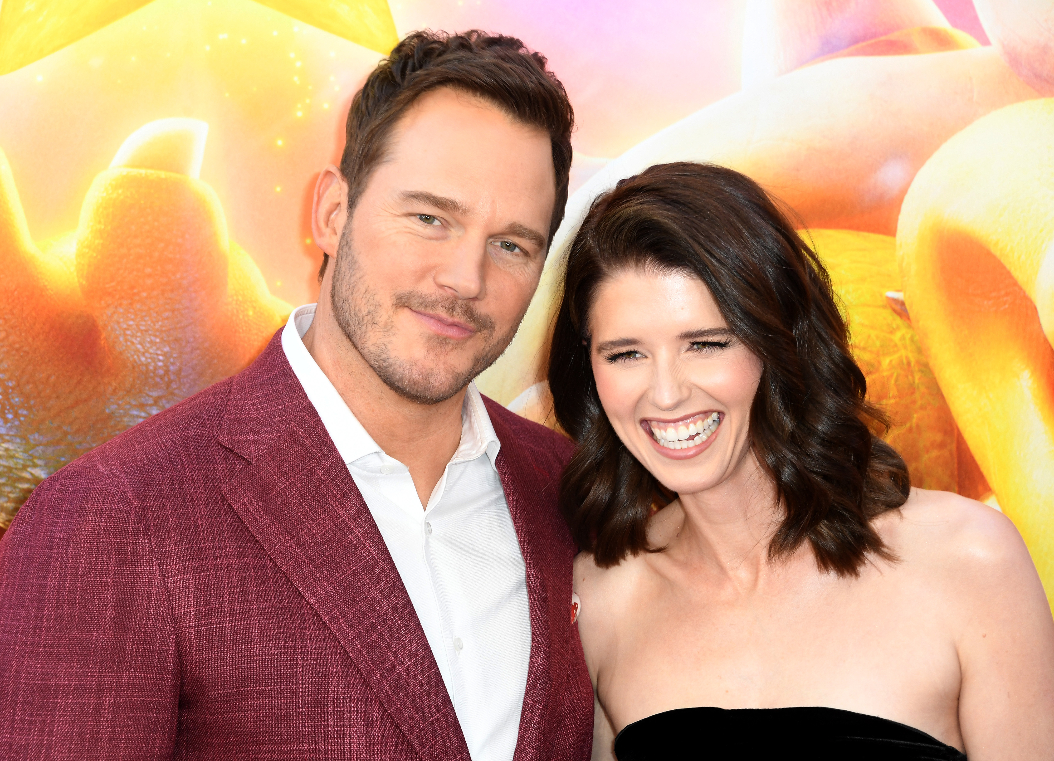 Chris Pratt in a burgundy blazer and white shirt poses with Katherine Schwarzenegger in a black strapless dress at a red carpet event, both smiling