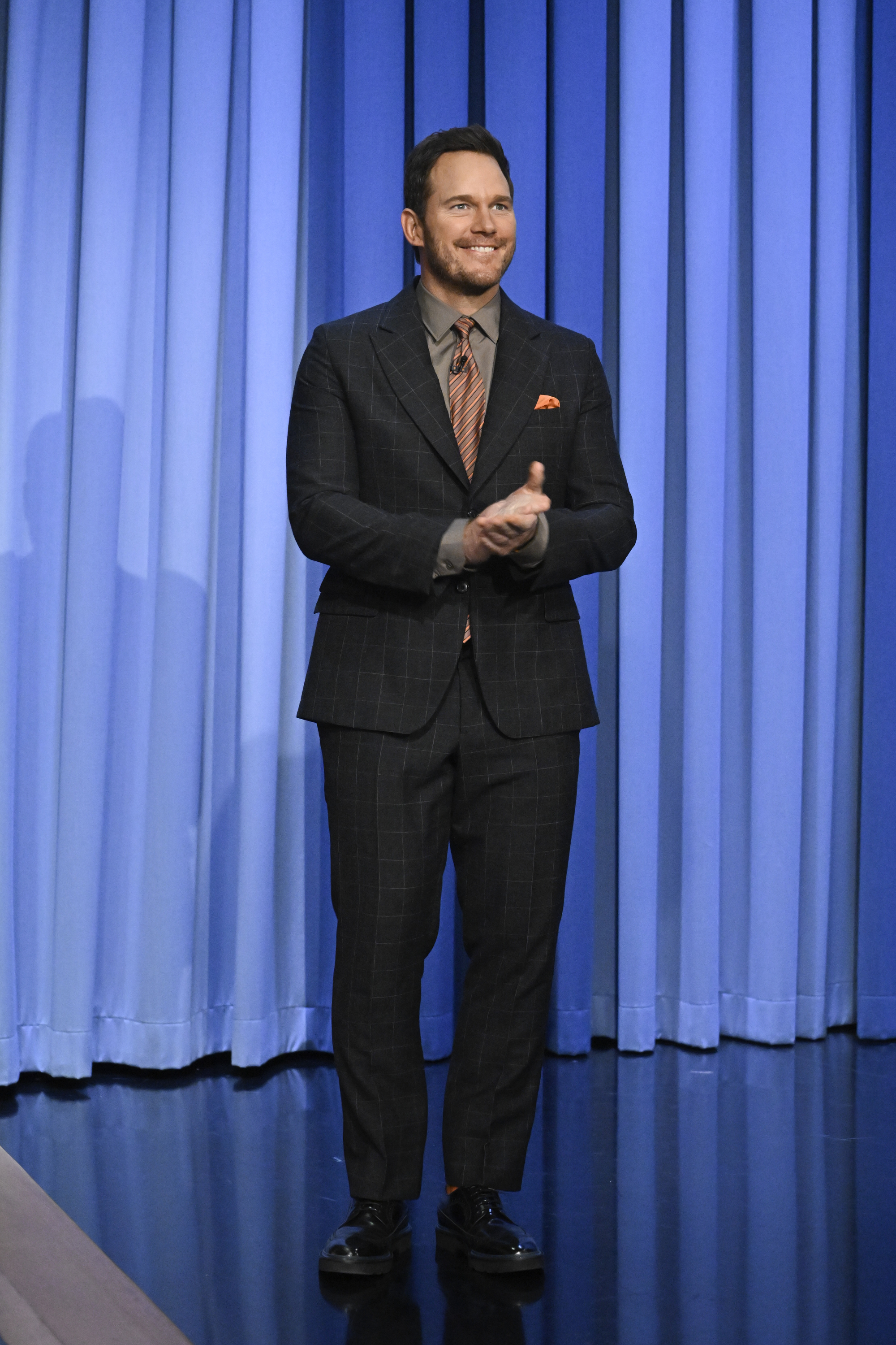 Chris Pratt standing and clapping while dressed in a dapper plaid suit and tie with a light pocket square, in front of a blue curtain background